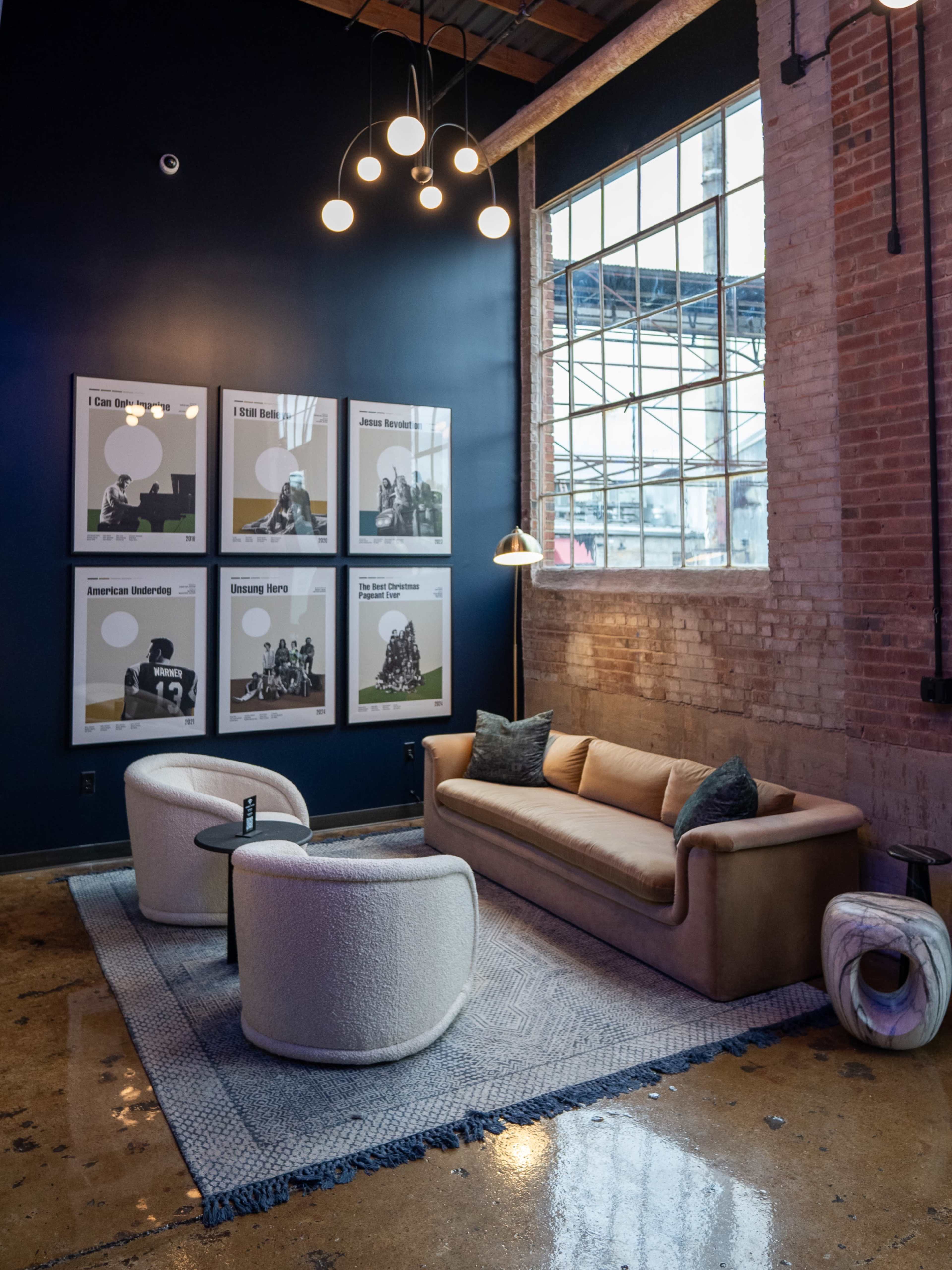 The image features a modern lounge area with a tan sofa, two round white chairs, and a large window surrounded by brick walls, adorned with several framed black-and-white artwork pieces.