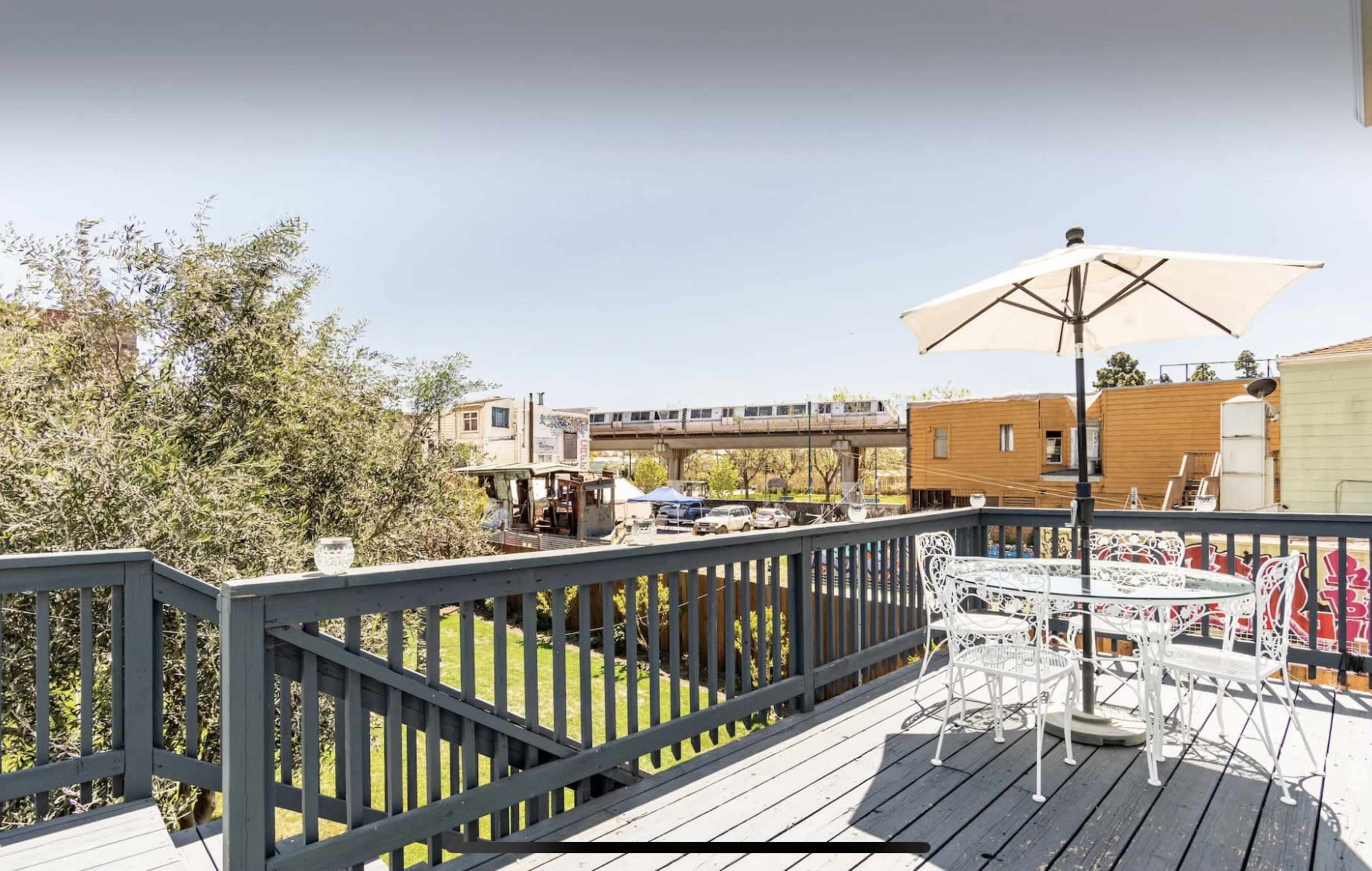 A wooden deck with a white table and chairs under an umbrella overlooks a neighborhood with buildings and a train passing nearby.