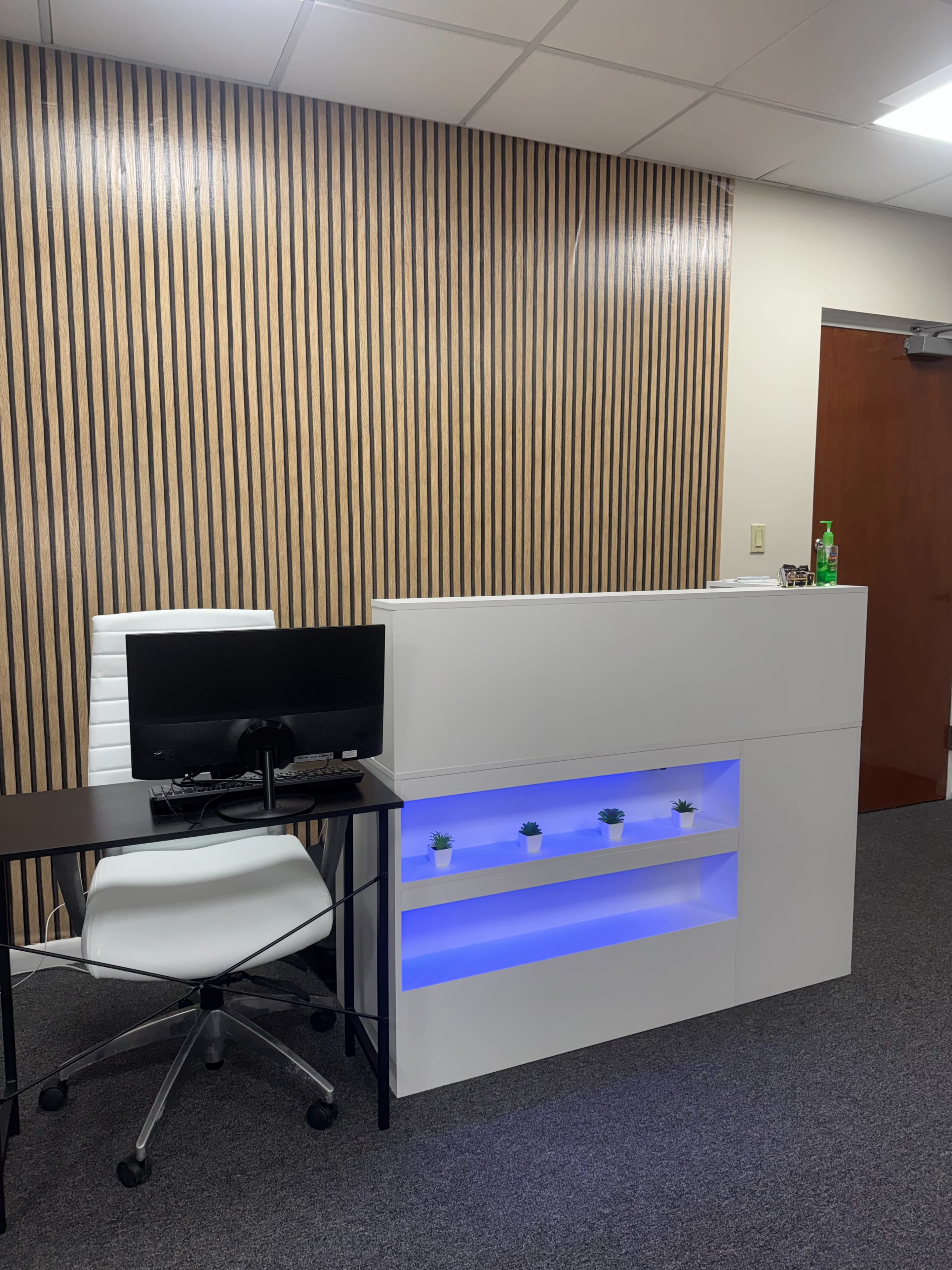 The image shows a modern reception area with a white desk featuring blue LED lighting, a computer monitor, a rolling chair, and a wooden wall behind it.