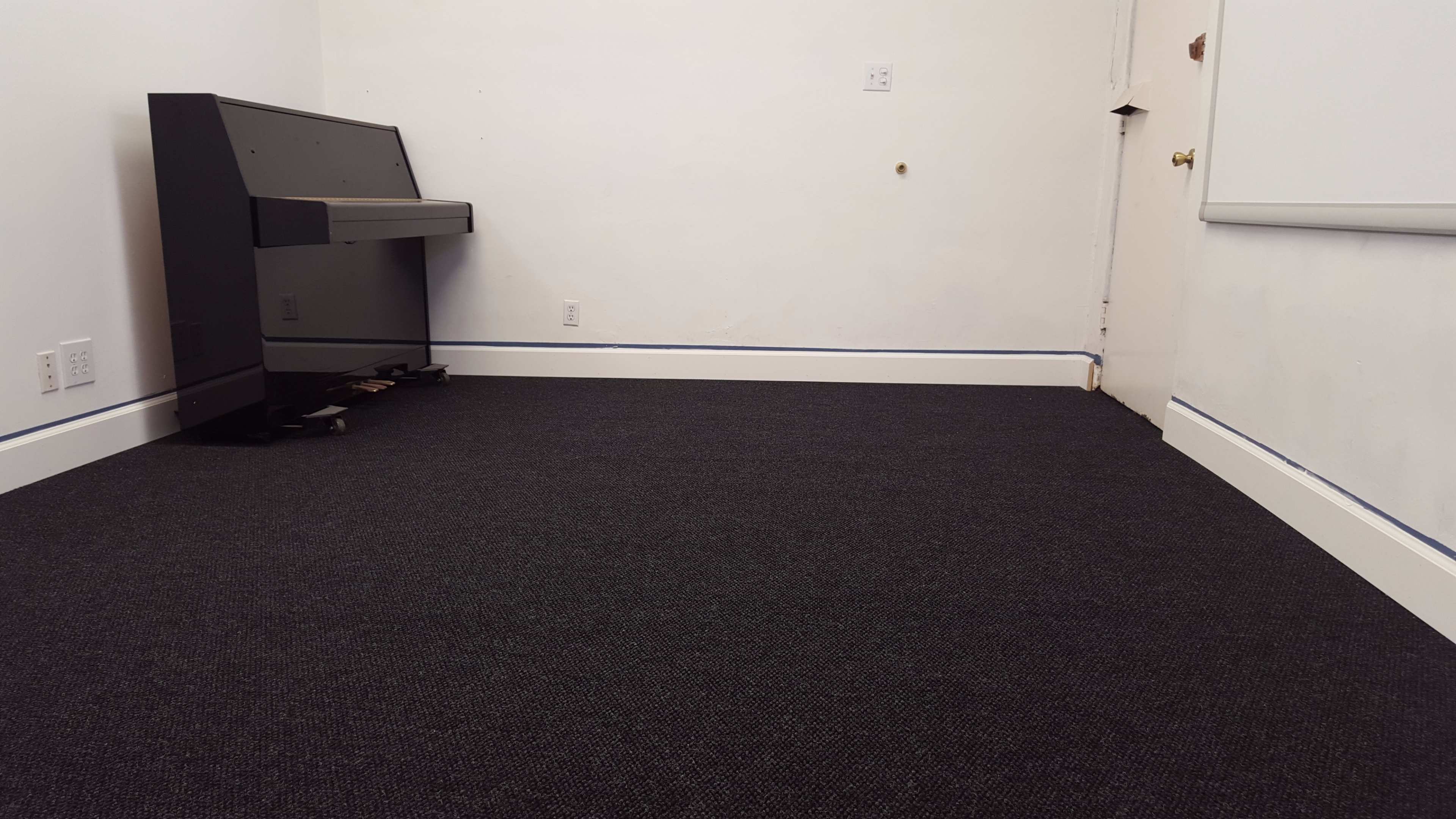 The image shows an empty room with dark carpet, a black piano in the corner, and a door on the right.