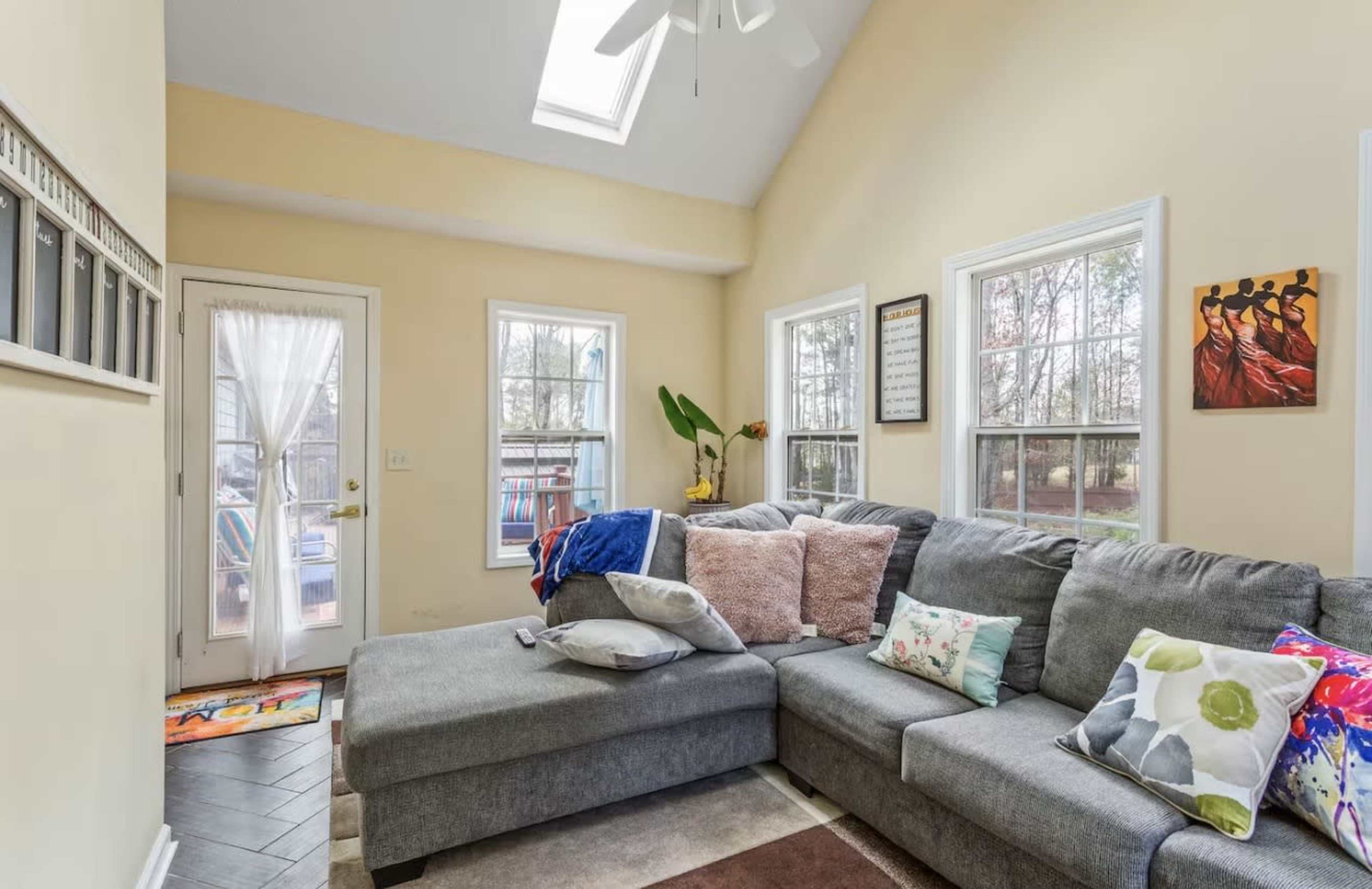 A spacious living room features a gray sectional sofa with colorful cushions, large windows, and a glass door leading to an outdoor area.