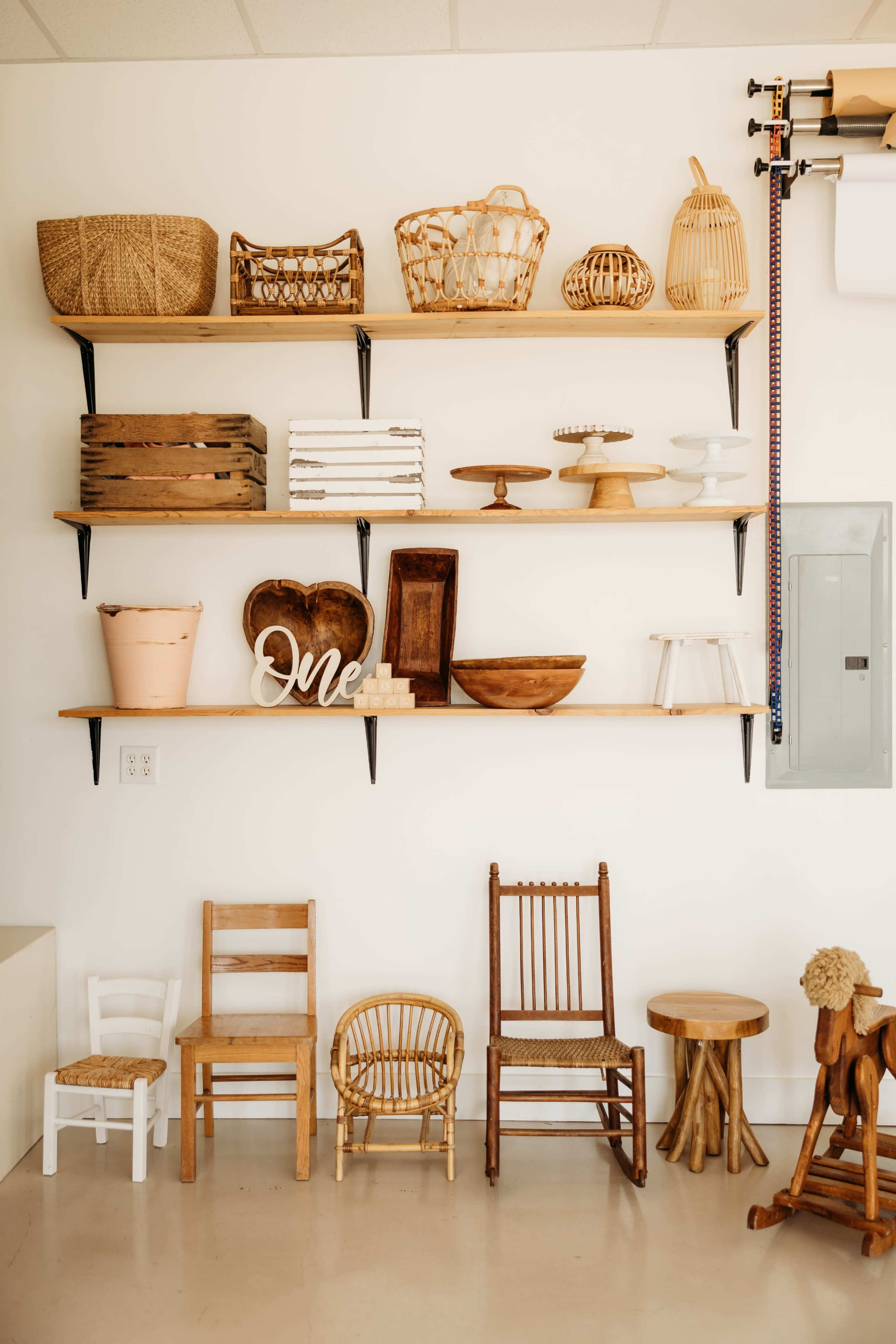 The image shows a wall with wooden shelves displaying various decorative items, including baskets and wooden bowls, above a collection of small wooden chairs.
