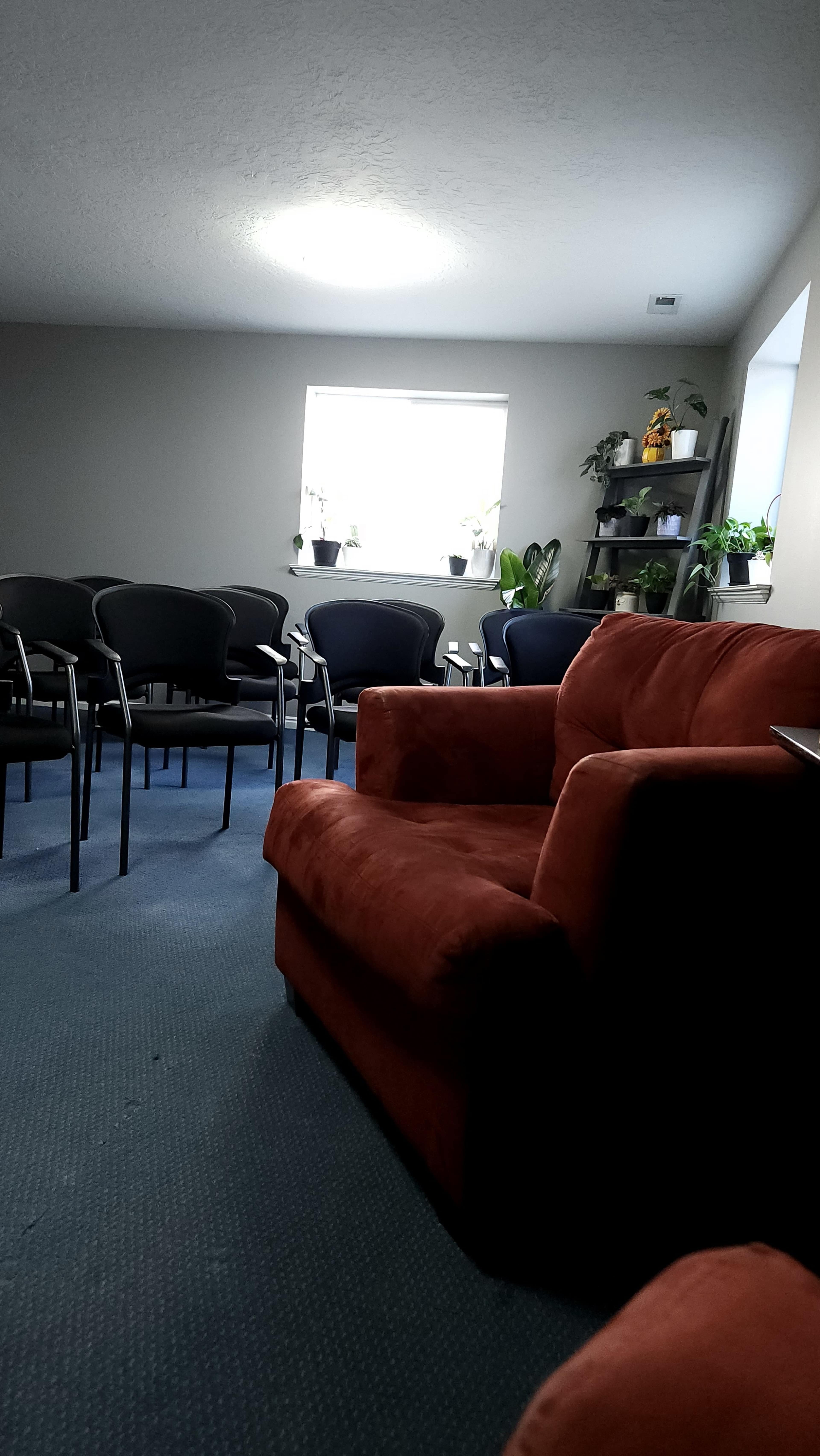 The image shows a waiting room featuring a red sofa, several black chairs arranged in rows, and potted plants on a shelf and windowsill.