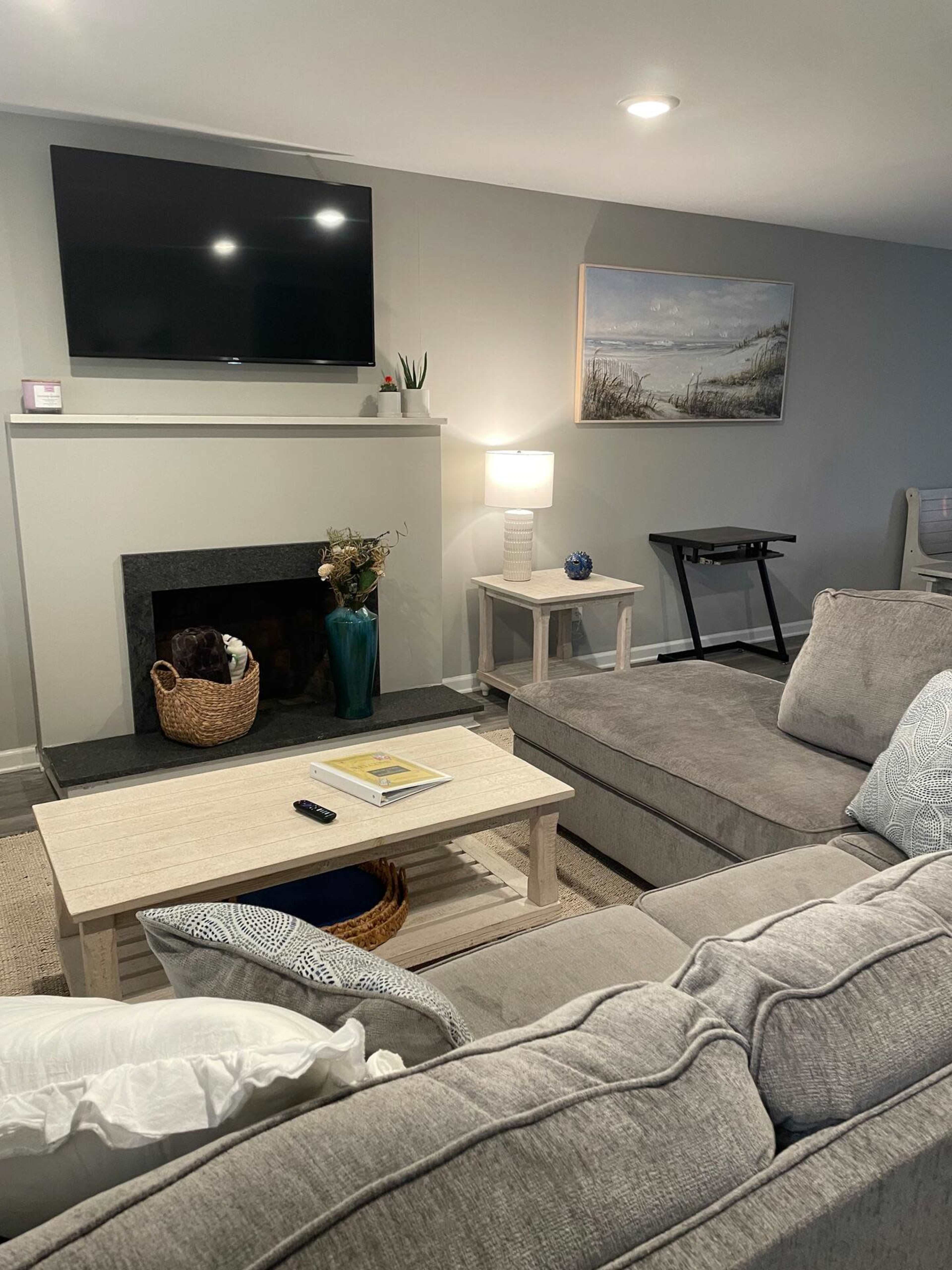 A cozy living room with a television mounted above a fireplace, a coffee table with a magazine, and a sofa arranged around a light-colored rug.