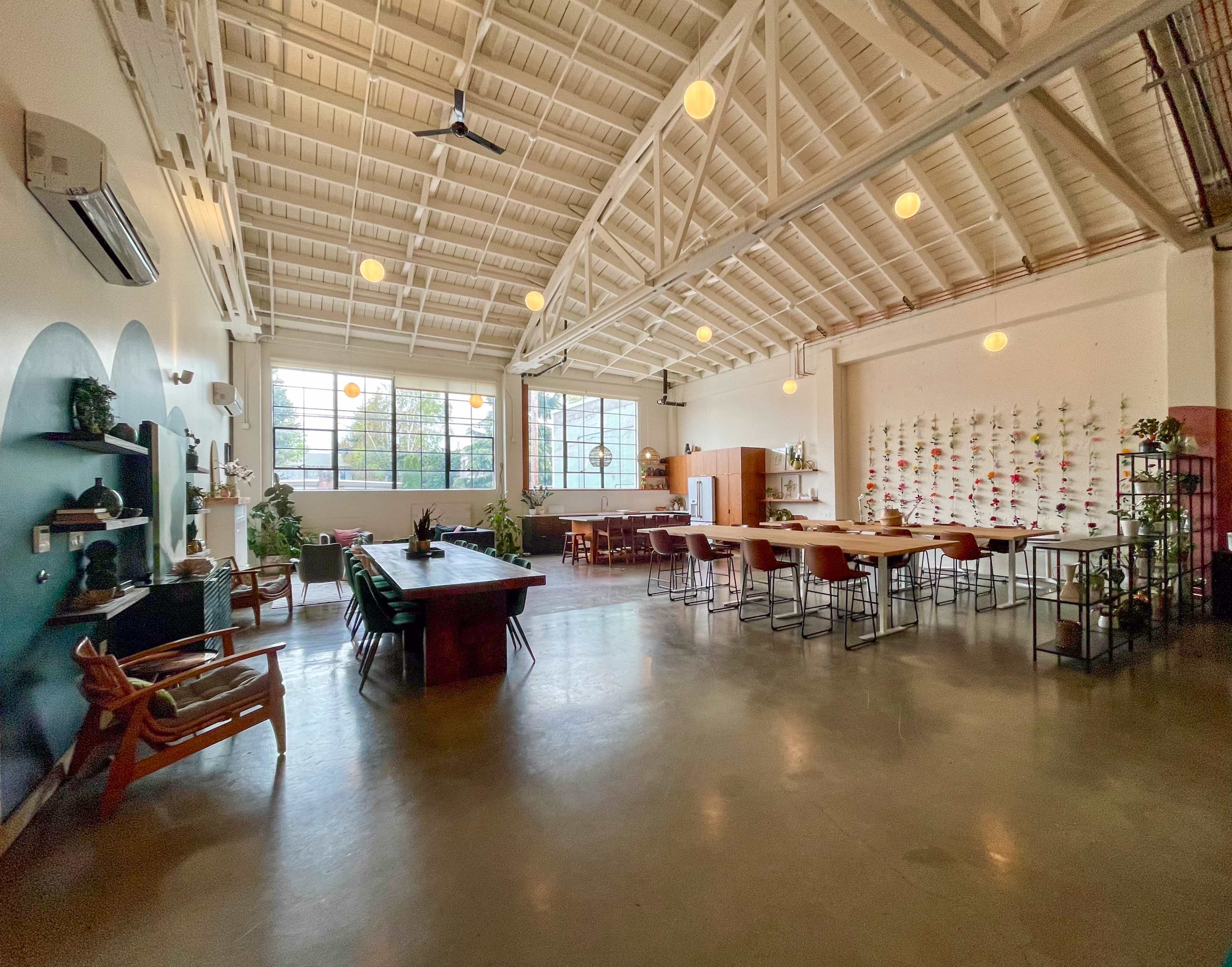 The image shows a spacious, bright interior of a dining or workspace with high ceilings, large windows, and various seating arrangements.