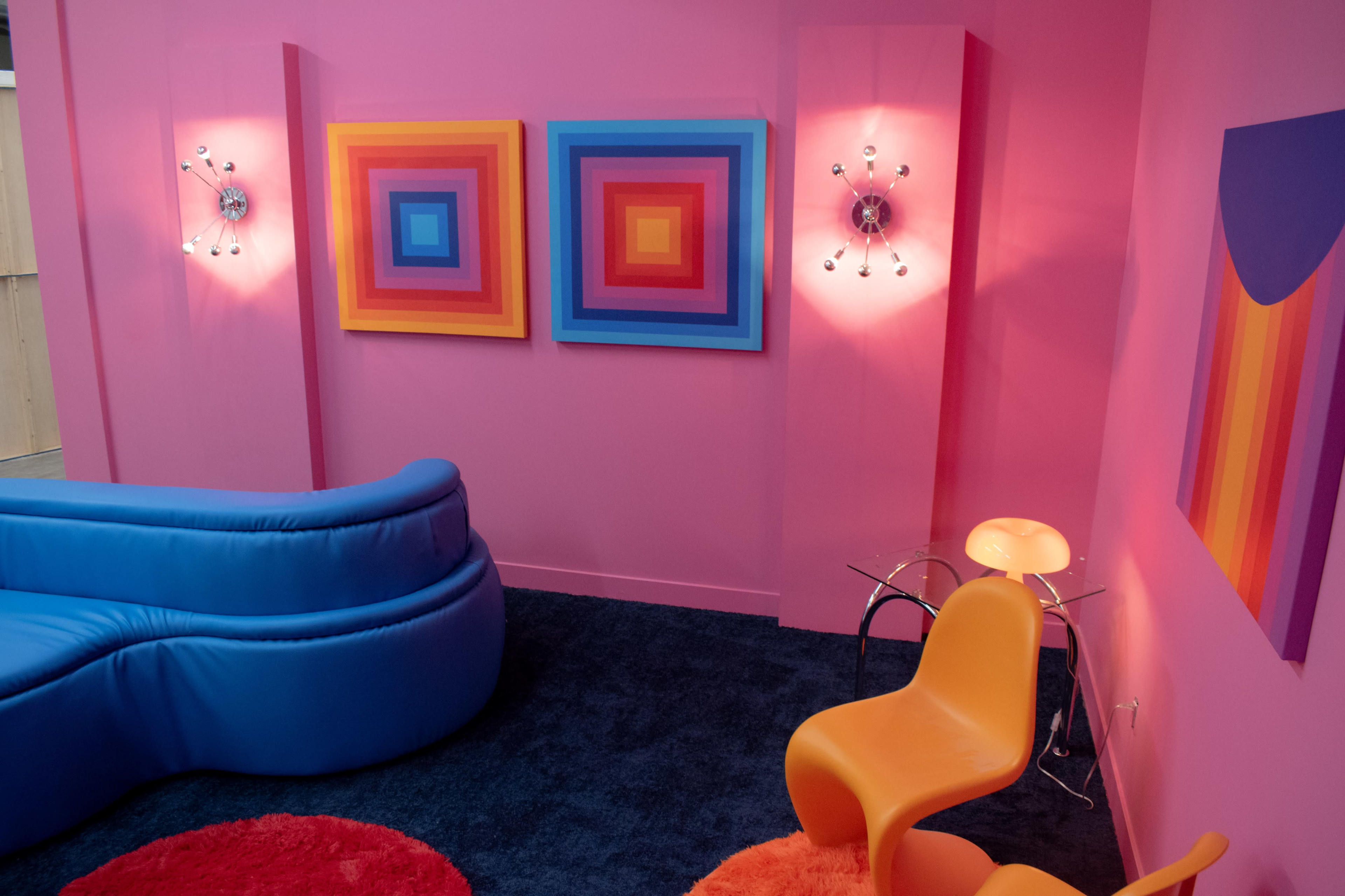 A vibrant room with pink walls, featuring two colorful abstract paintings and mid-century modern furniture, including a blue curved sofa and orange chairs.