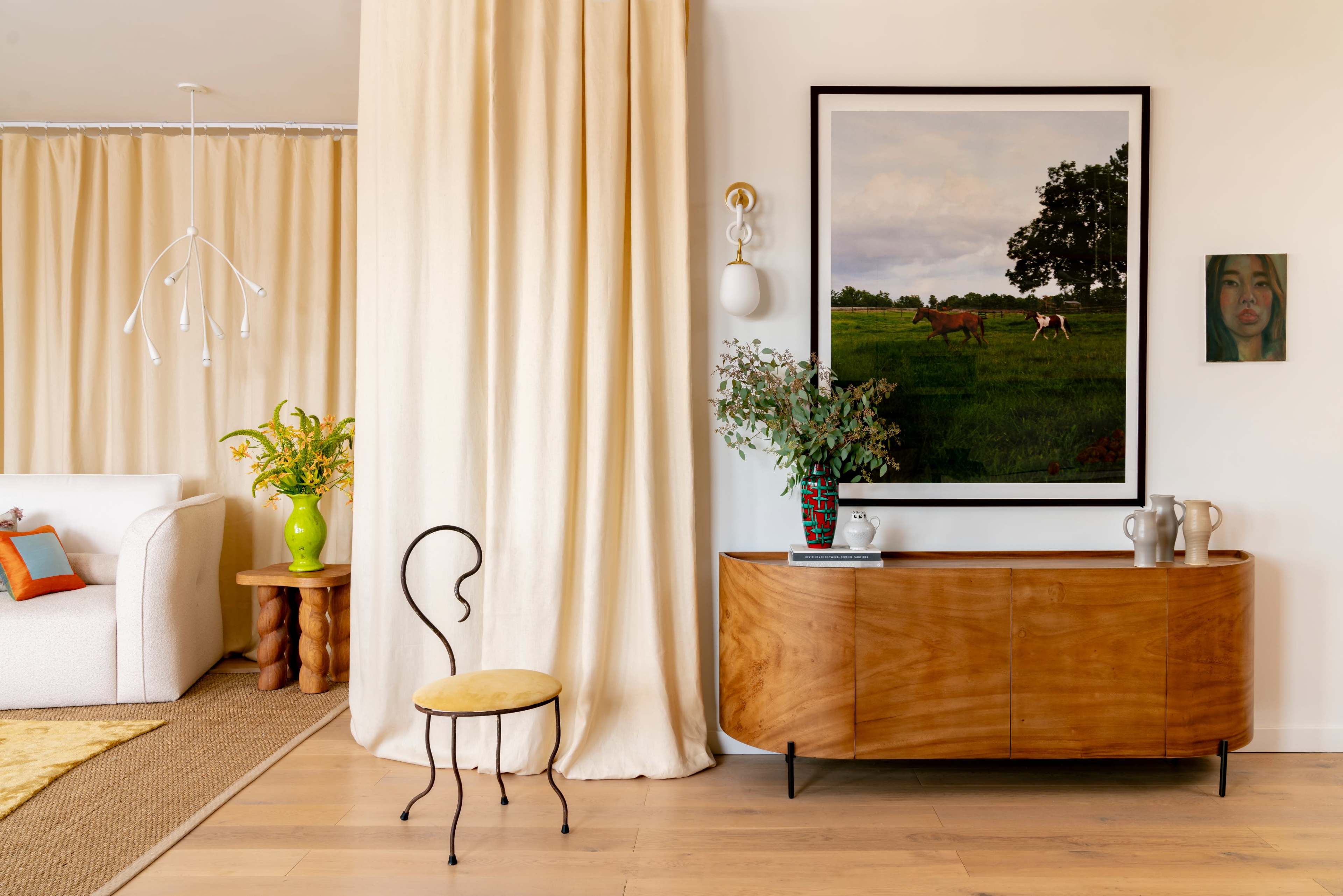 The scene features a modern living space with a wooden sideboard, a chair, a large framed landscape painting of horses, and decorative elements including a vase with flowers and a portrait on the wall.