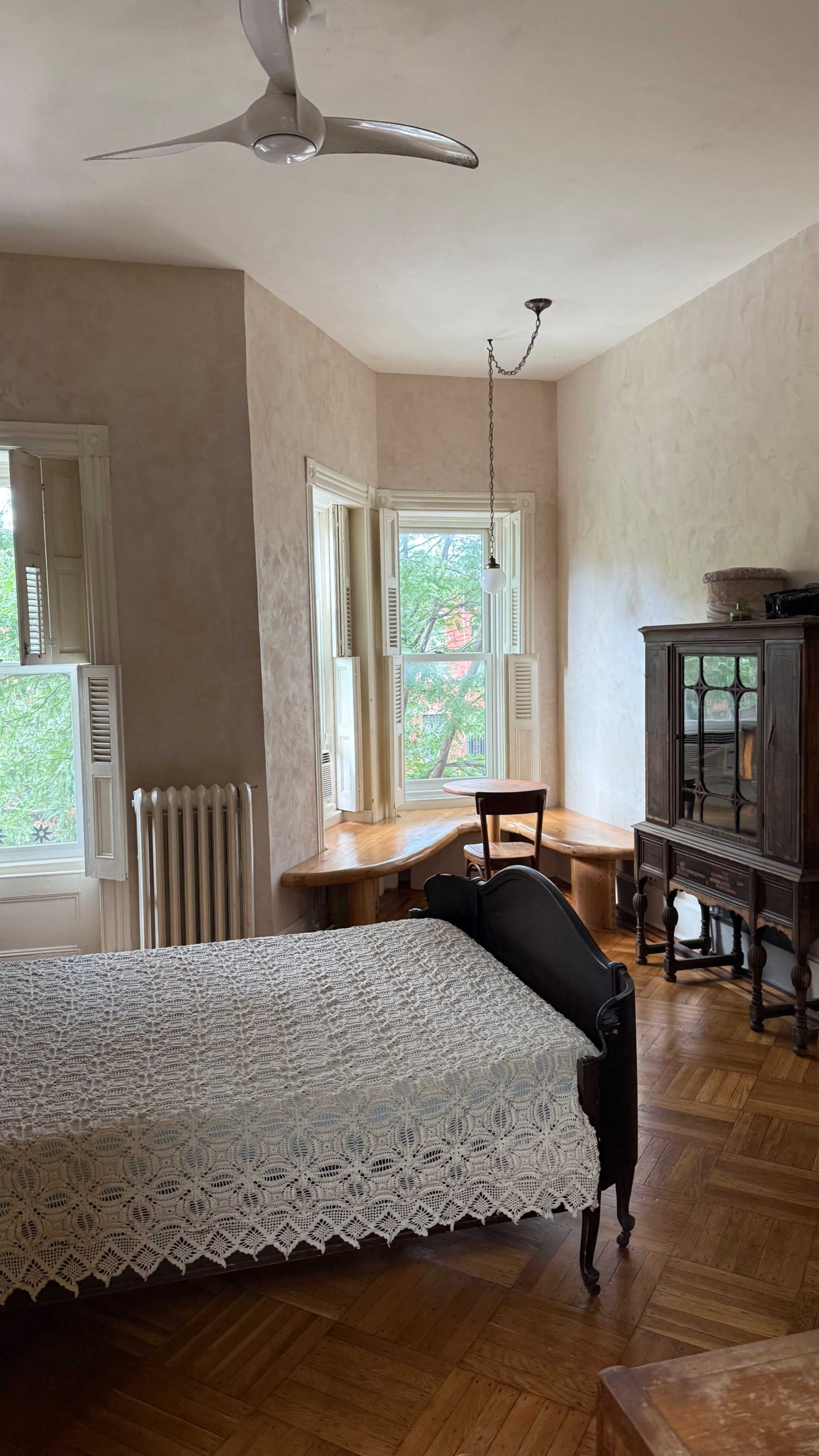 A bedroom features a wooden bed, a small table by a window, and an antique cabinet against a wall.