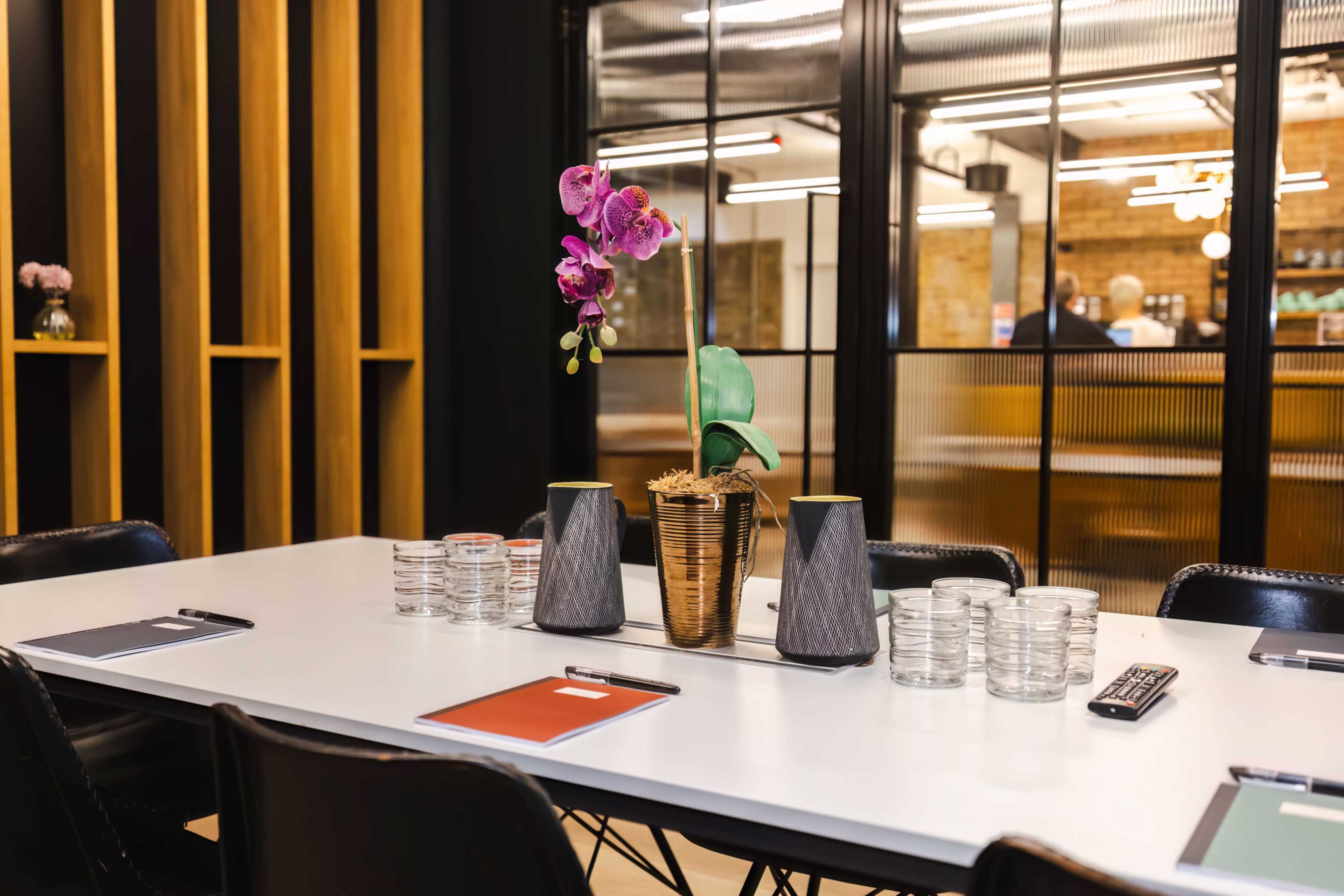 A modern meeting room features a white table set with glassware, colorful notepads, and a decorative orchid in a gold and marble vase.