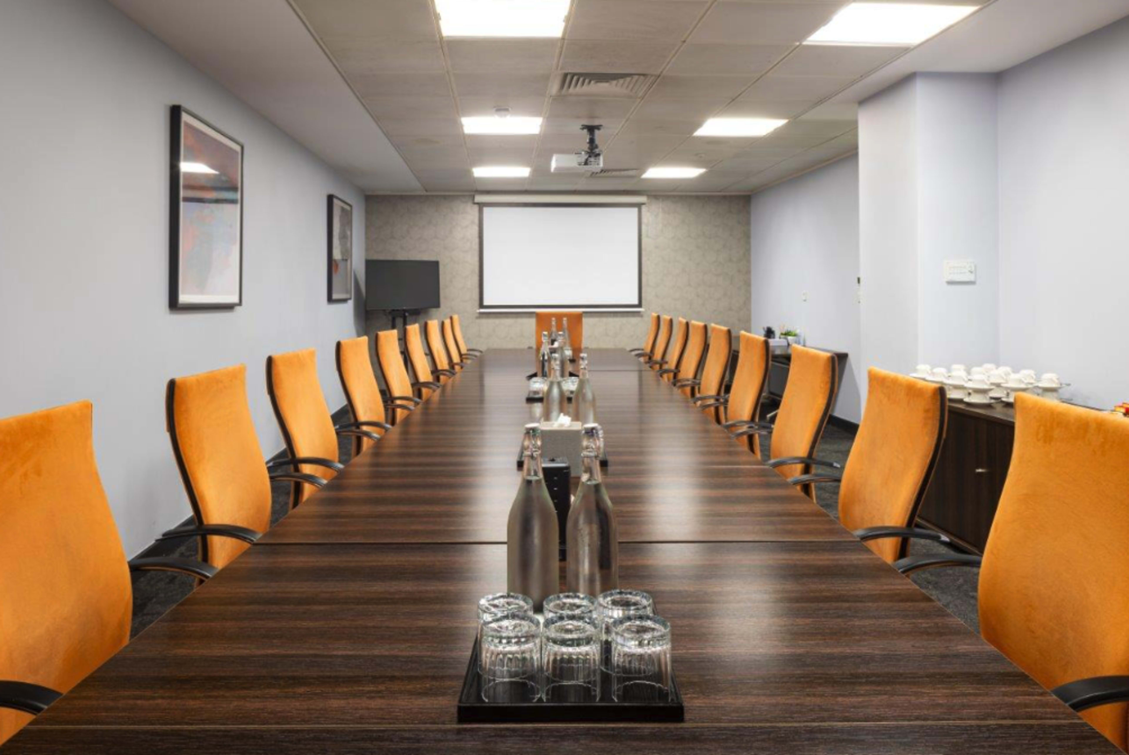 A long conference table with orange chairs is set up in a meeting room, featuring drinking glasses and water bottles, with a projector and screen at one end.