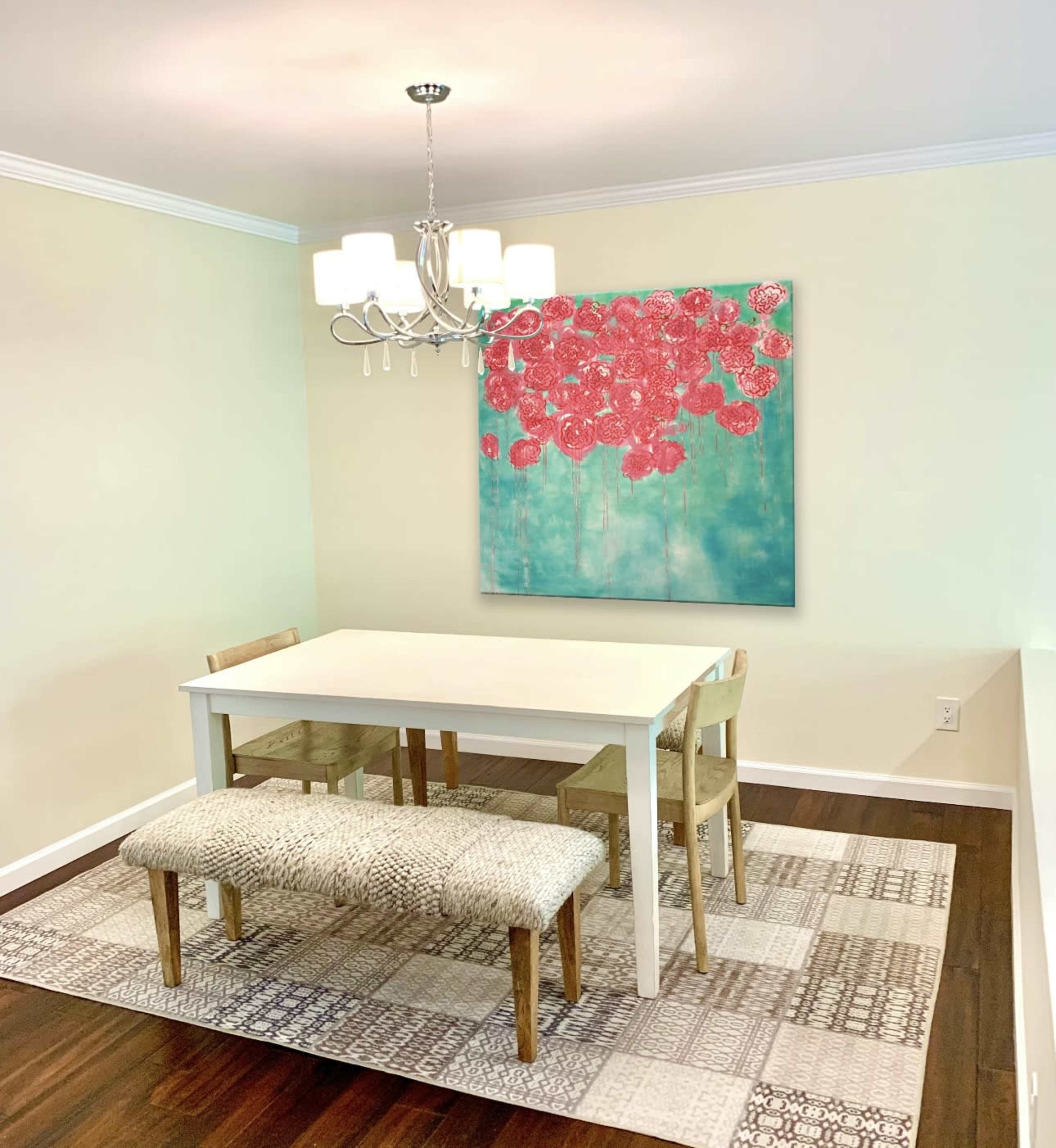 A dining area features a white table and two wooden chairs, with a bench and a large floral painting on the wall above, illuminated by a chandelier.
