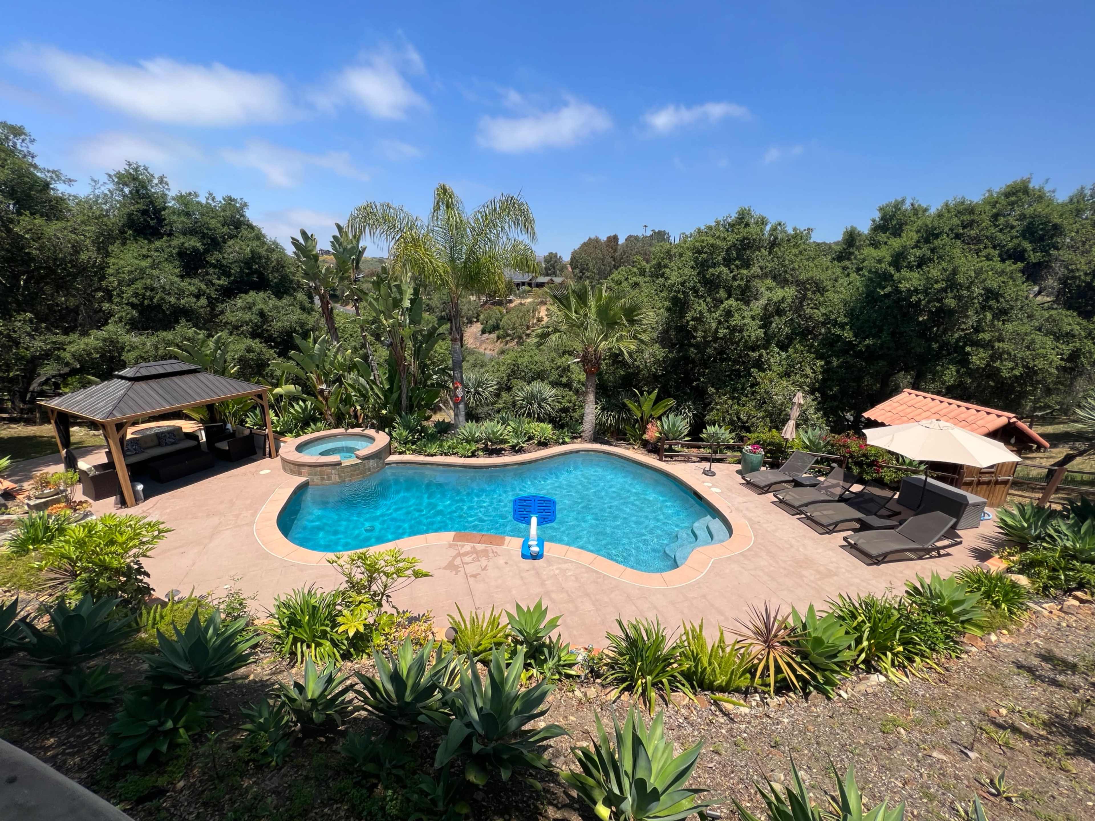 A backyard area with a swimming pool, hot tub, lounge chairs, and a gazebo surrounded by lush greenery.