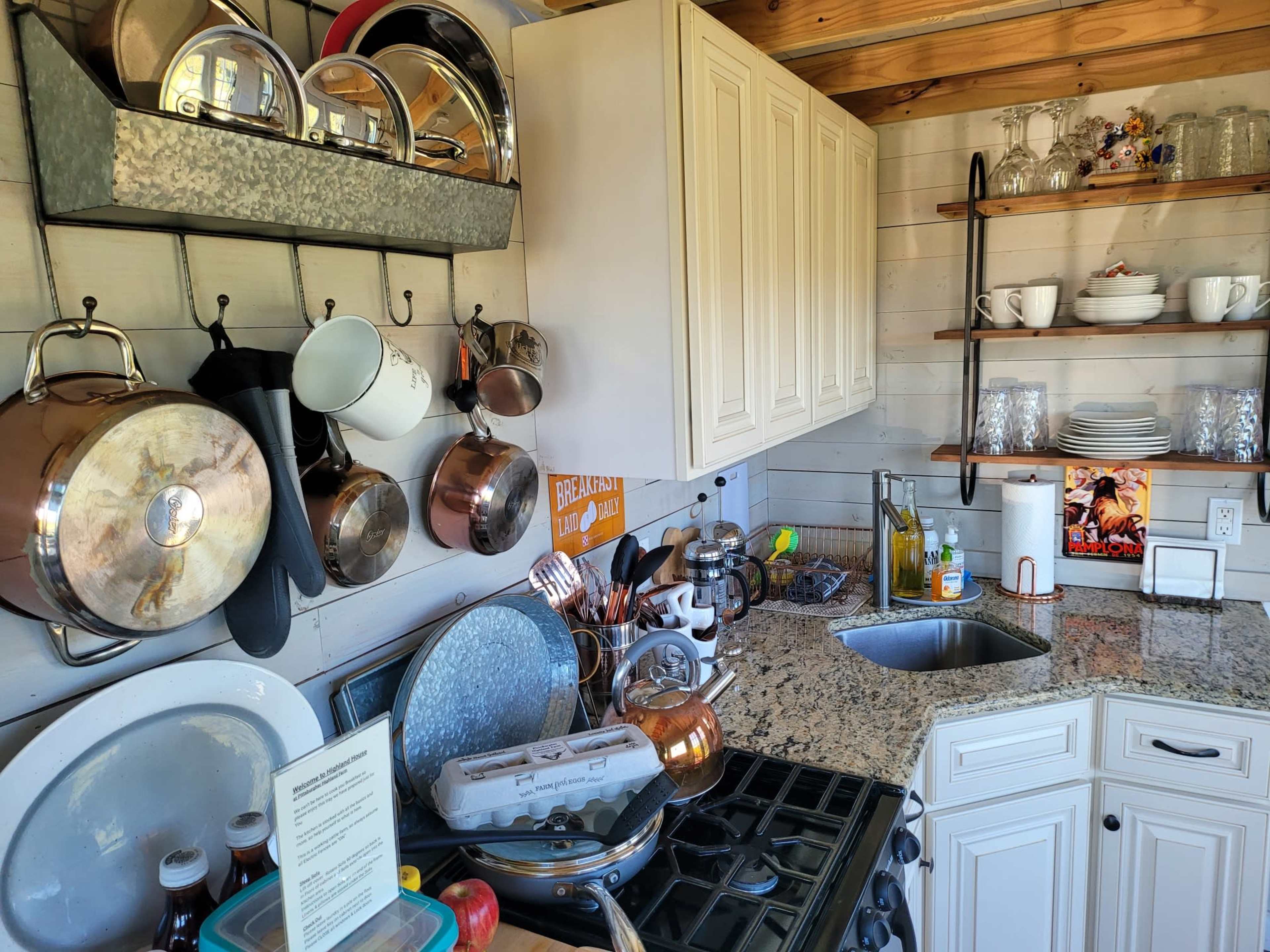 A kitchen with metal pots and pans hanging on the wall, a granite countertop, and dishes organized on shelves.