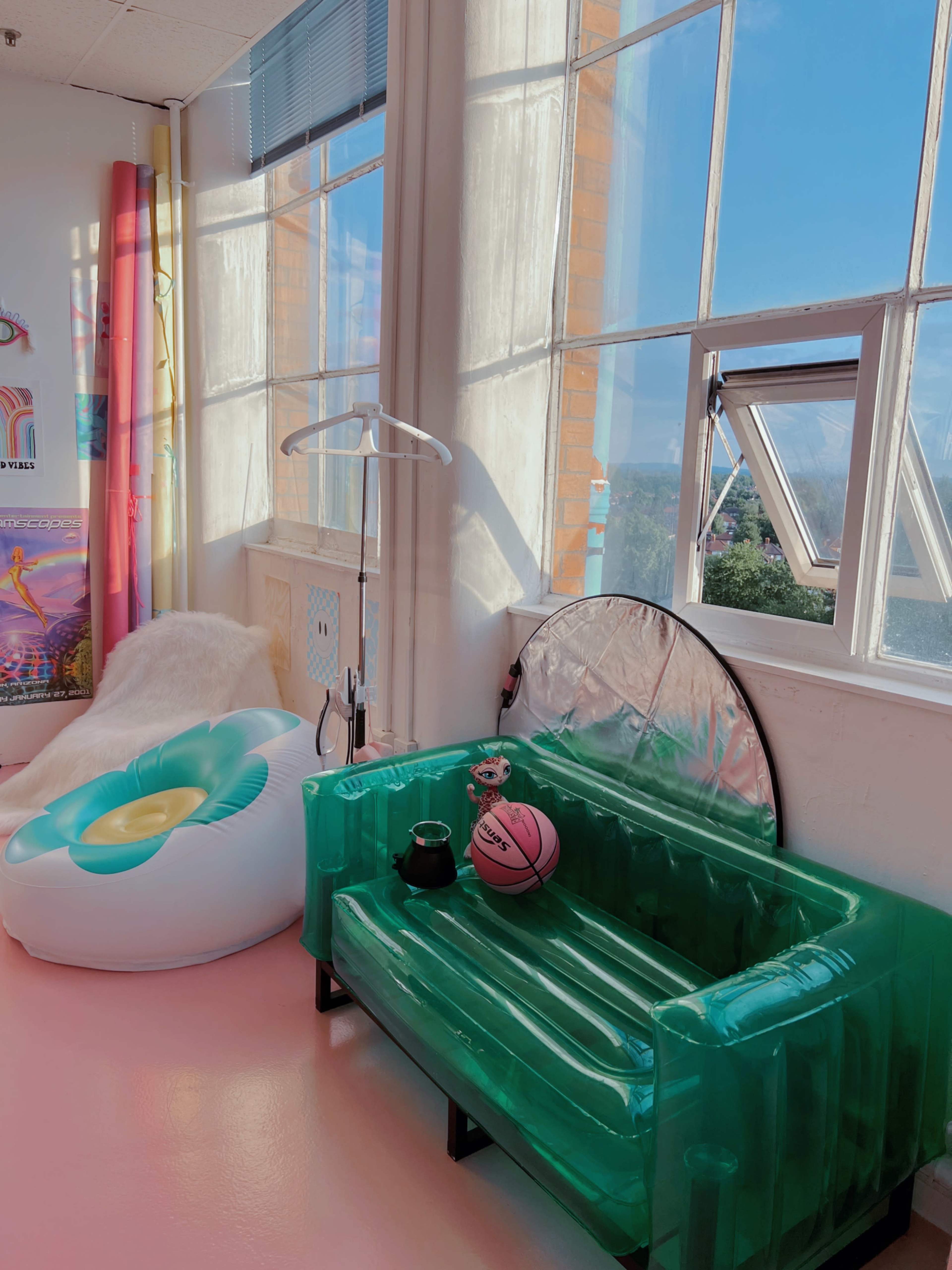 A brightly lit room with large windows, featuring a green inflatable couch, a white bean bag chair, a basketball, and various decorative elements.
