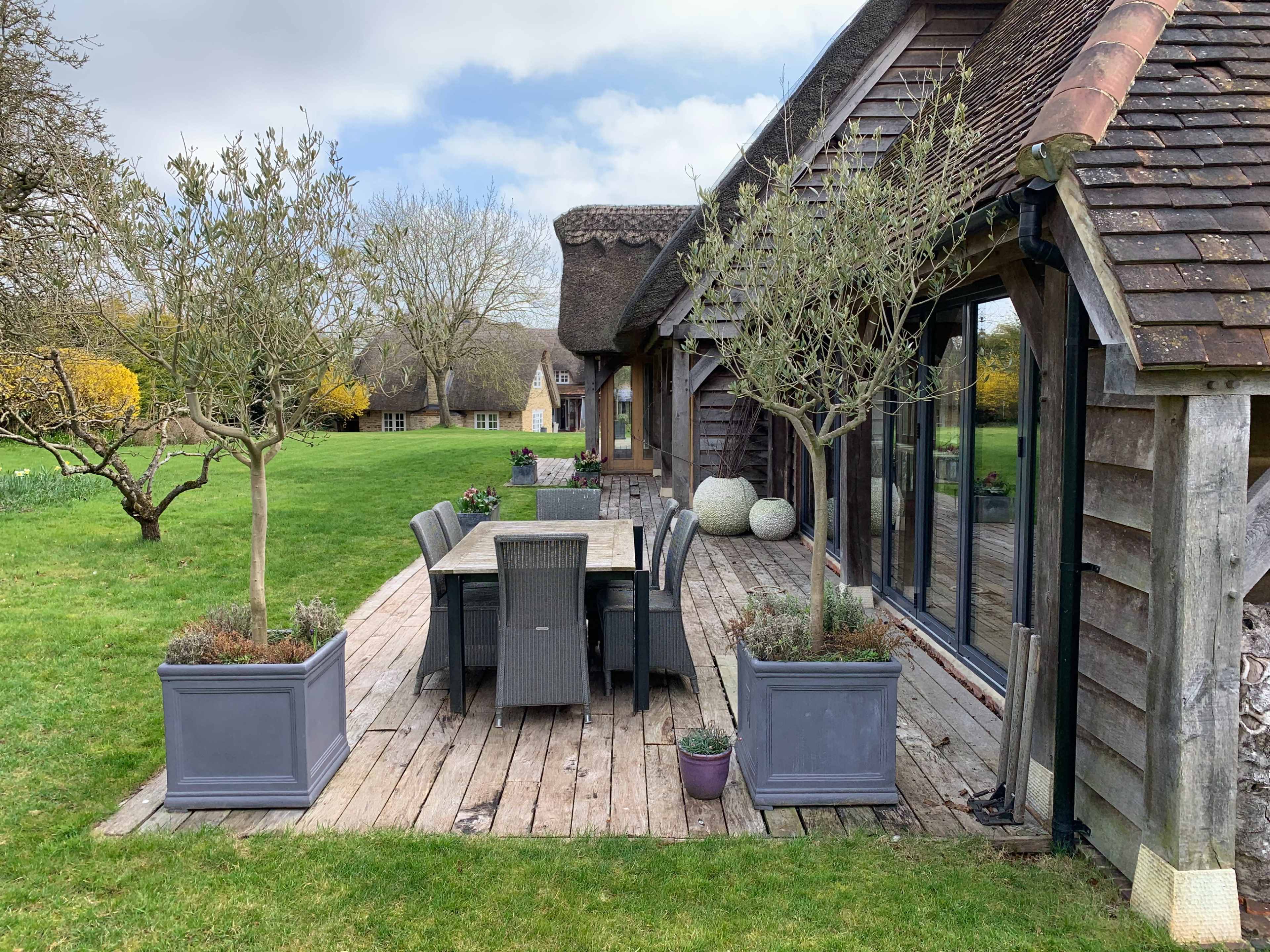 A wooden deck with a dining table and chairs is surrounded by potted plants and trees beside a house with a thatched roof.