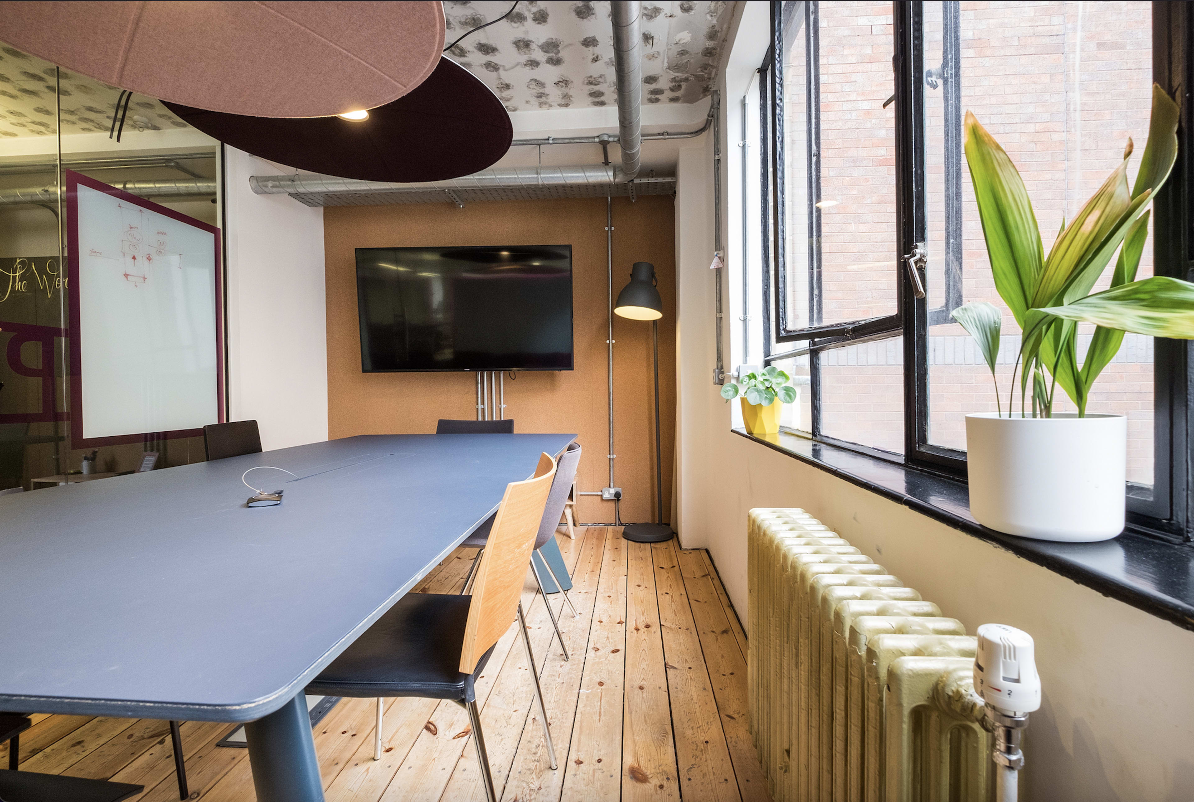A modern conference room features a long table, several wooden chairs, a wall-mounted television, and a window with a potted plant beside a radiator.