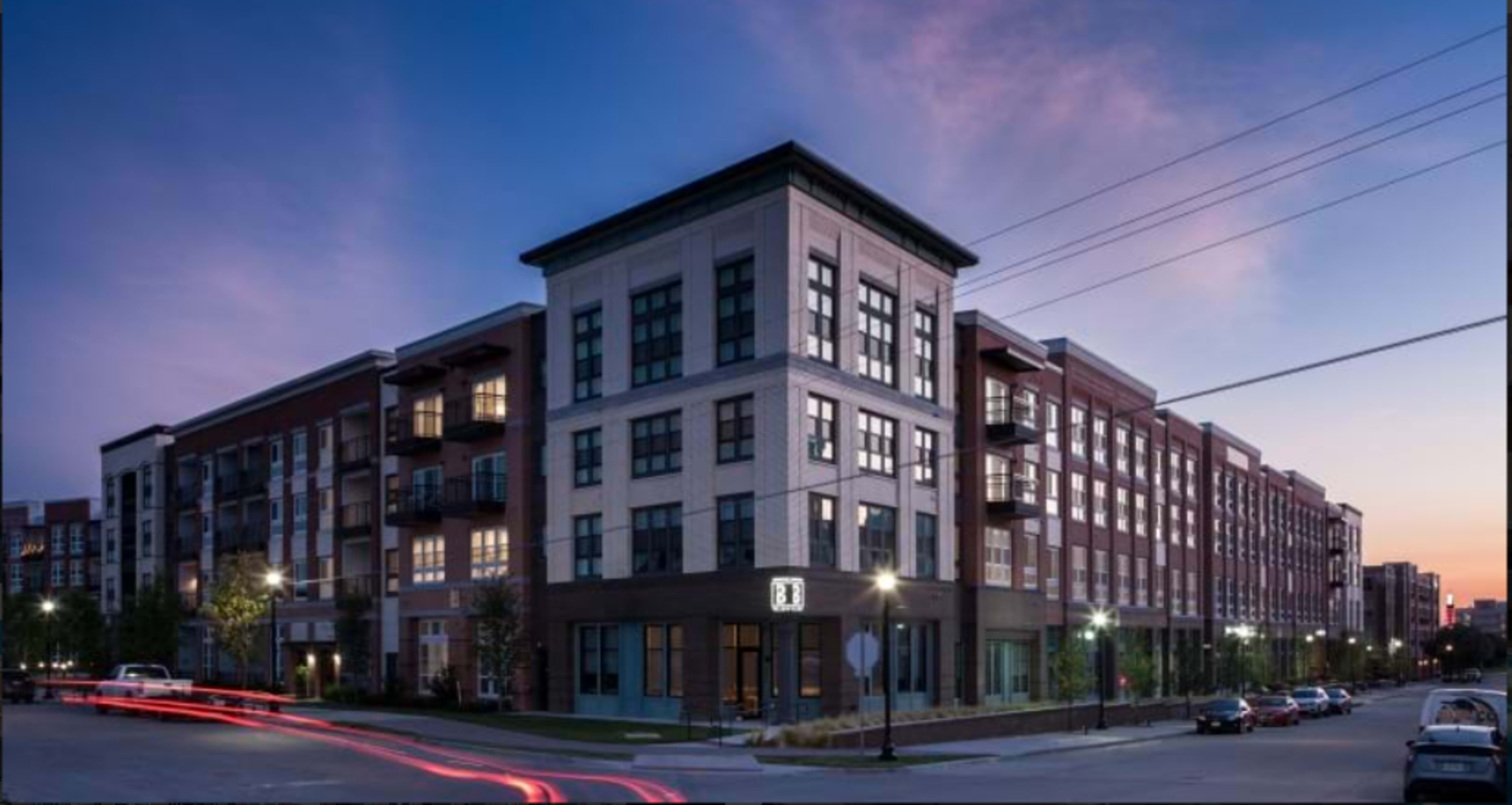 A modern apartment building with several stories and large windows is illuminated at dusk, with light trails from passing cars visible in the foreground.
