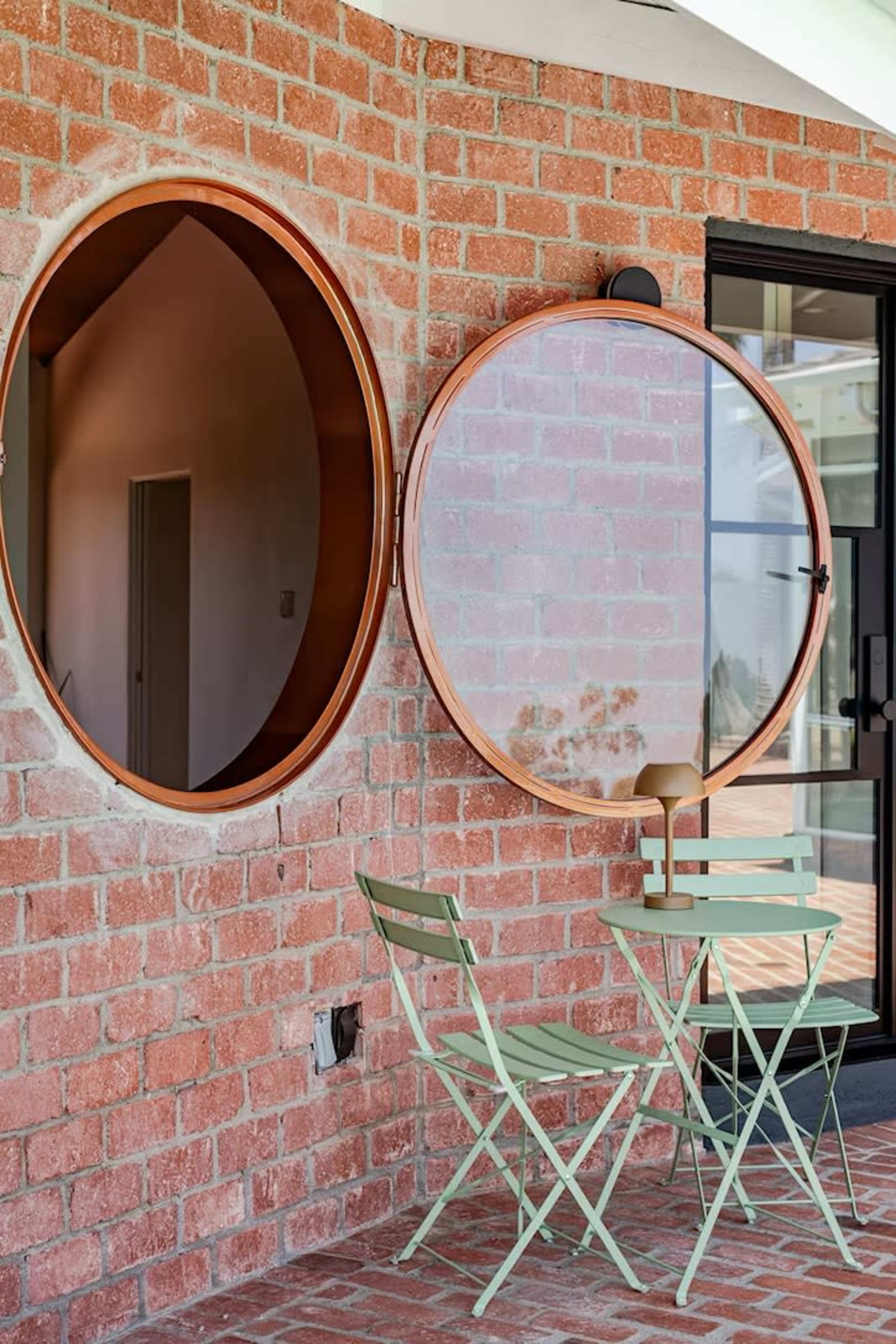 The image shows two circular mirrors mounted on a brick wall beside a small green table and chairs set on a patio.