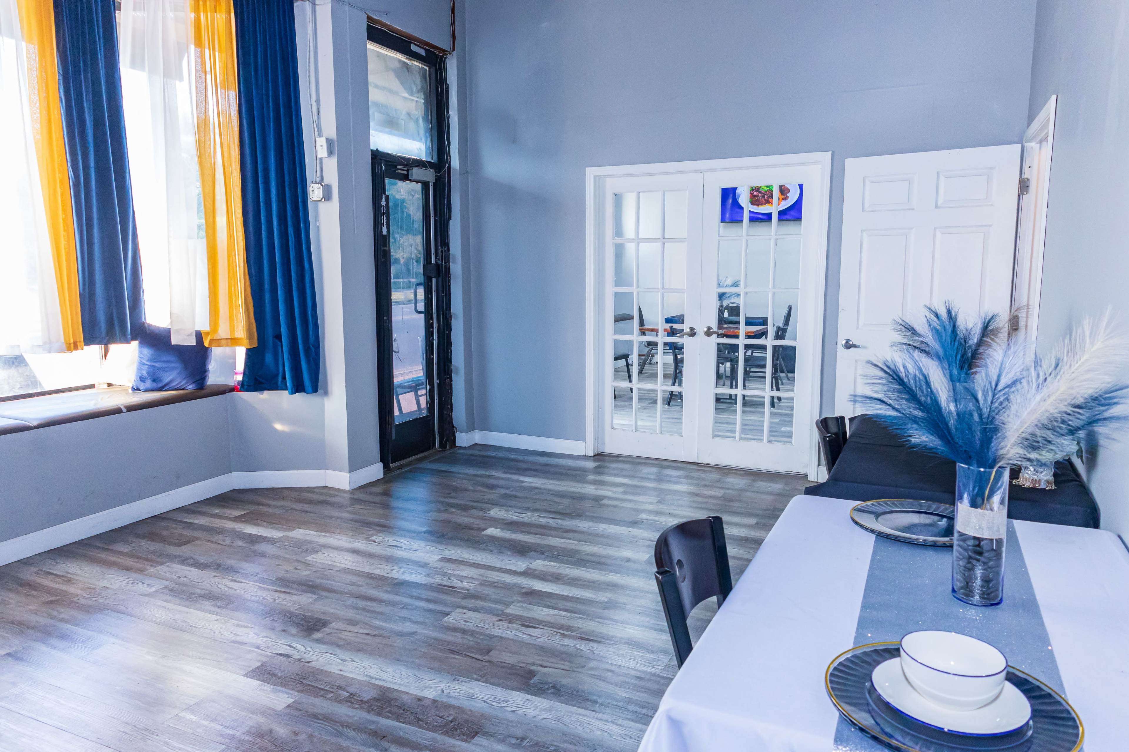 The image shows a bright, open room with large windows dressed in orange and blue curtains, a table set with dishes, and a door leading to another space.