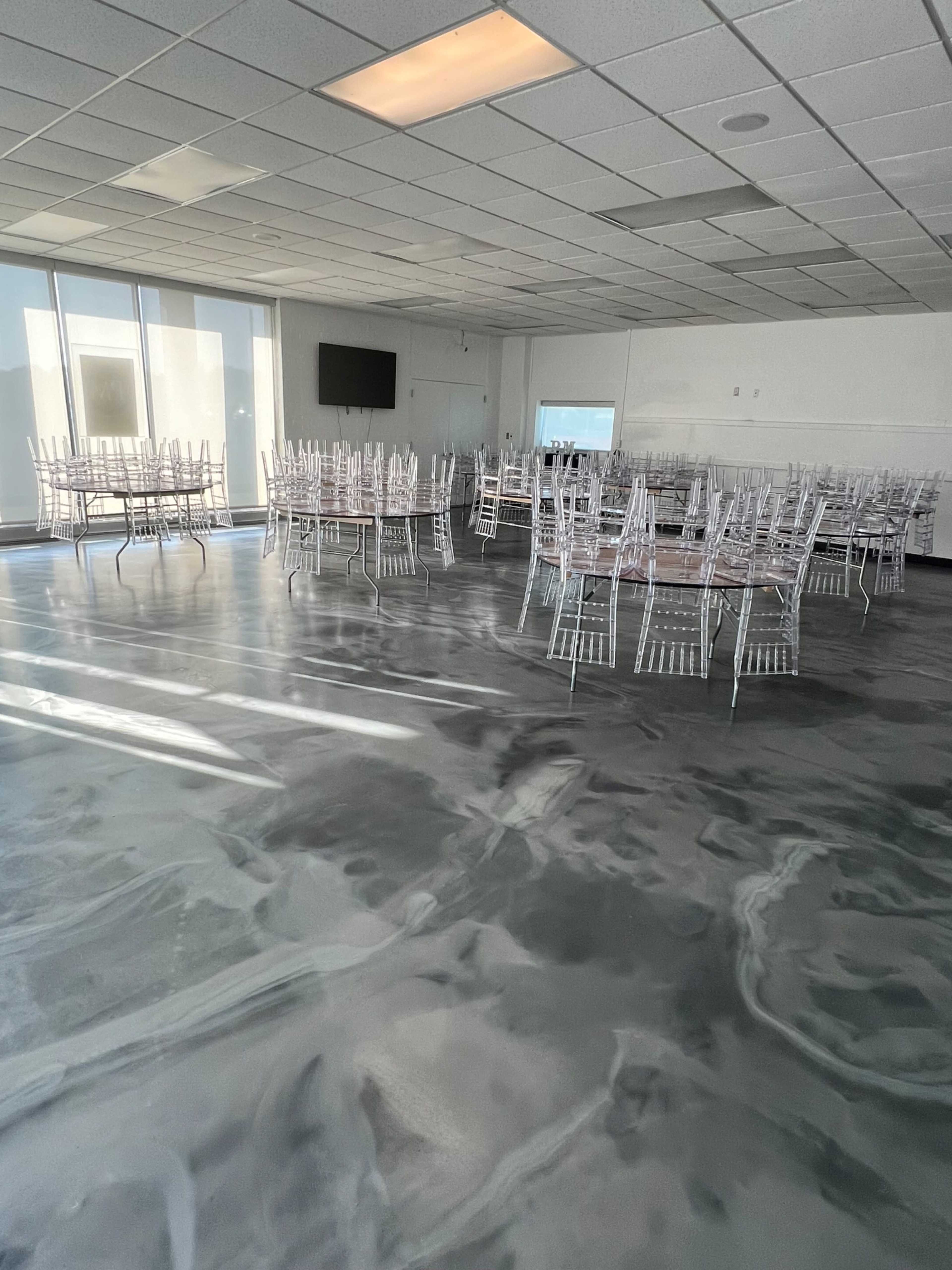 A spacious room with several round tables set up, each surrounded by transparent chiavari chairs, on a polished floor.
