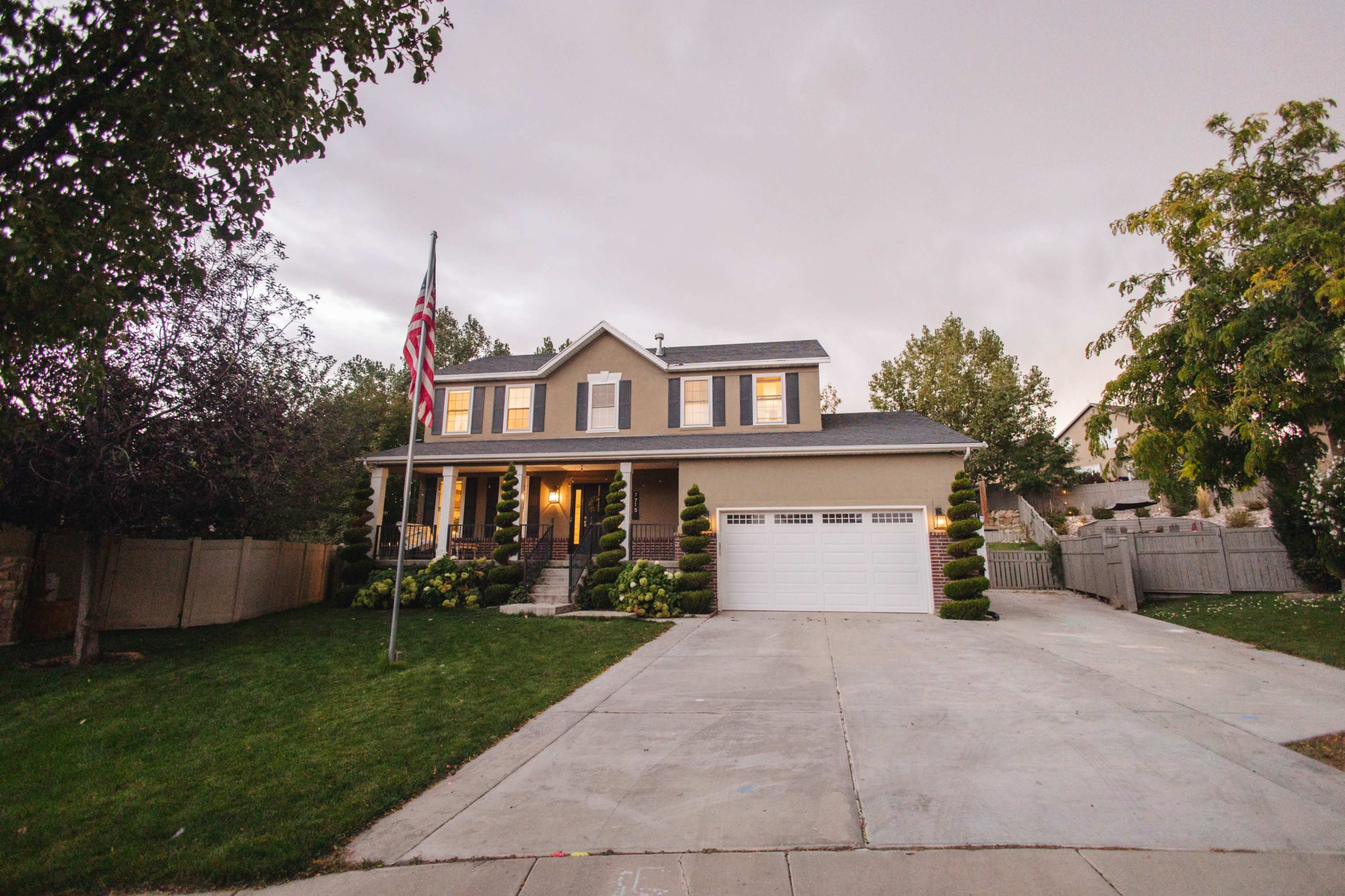A two-story house with a front porch, American flag, and neatly landscaped yard is situated on a paved driveway.
