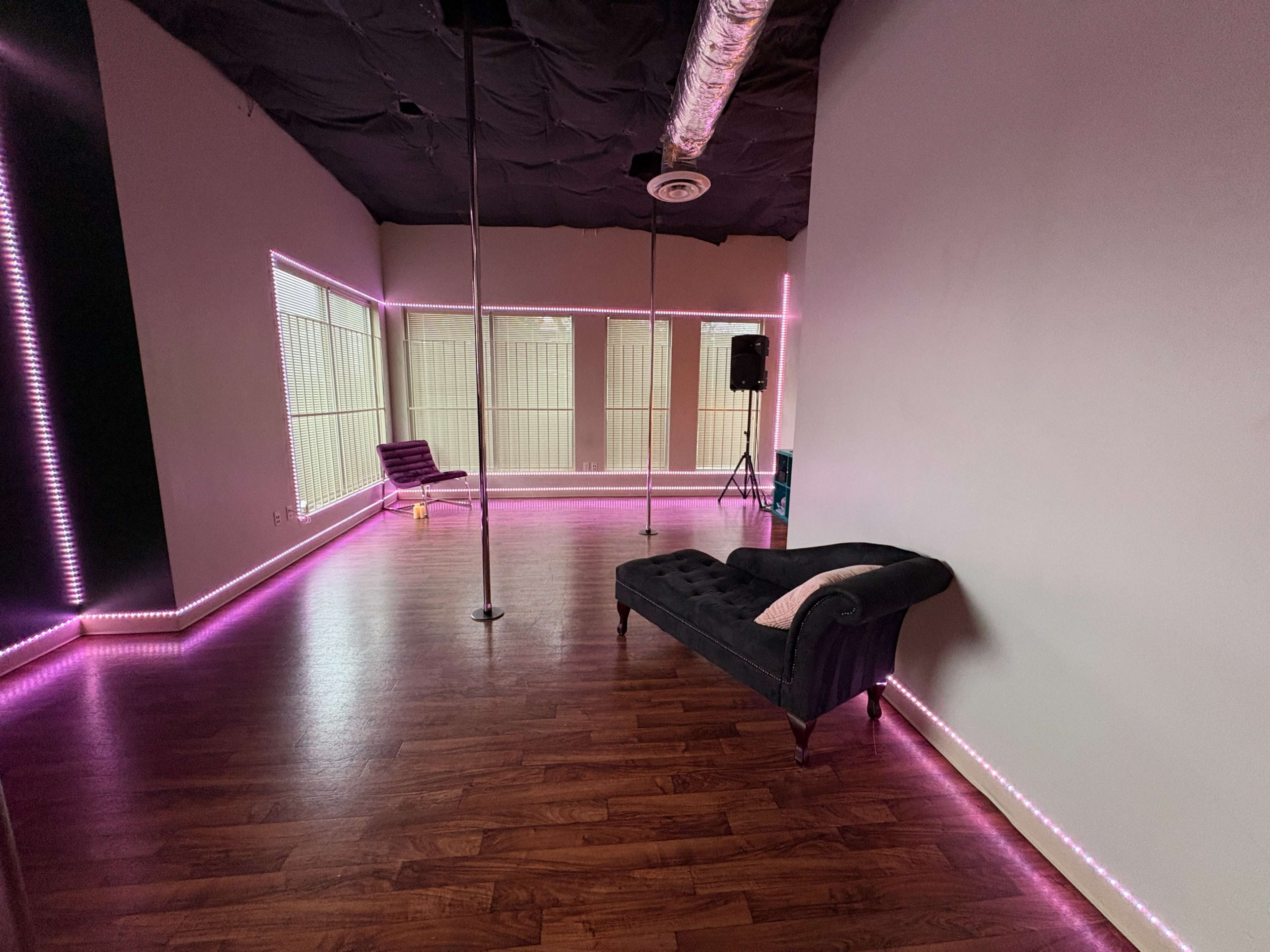 The image shows a dance studio featuring a pole, a chaise lounge, and pink LED lighting along the walls.