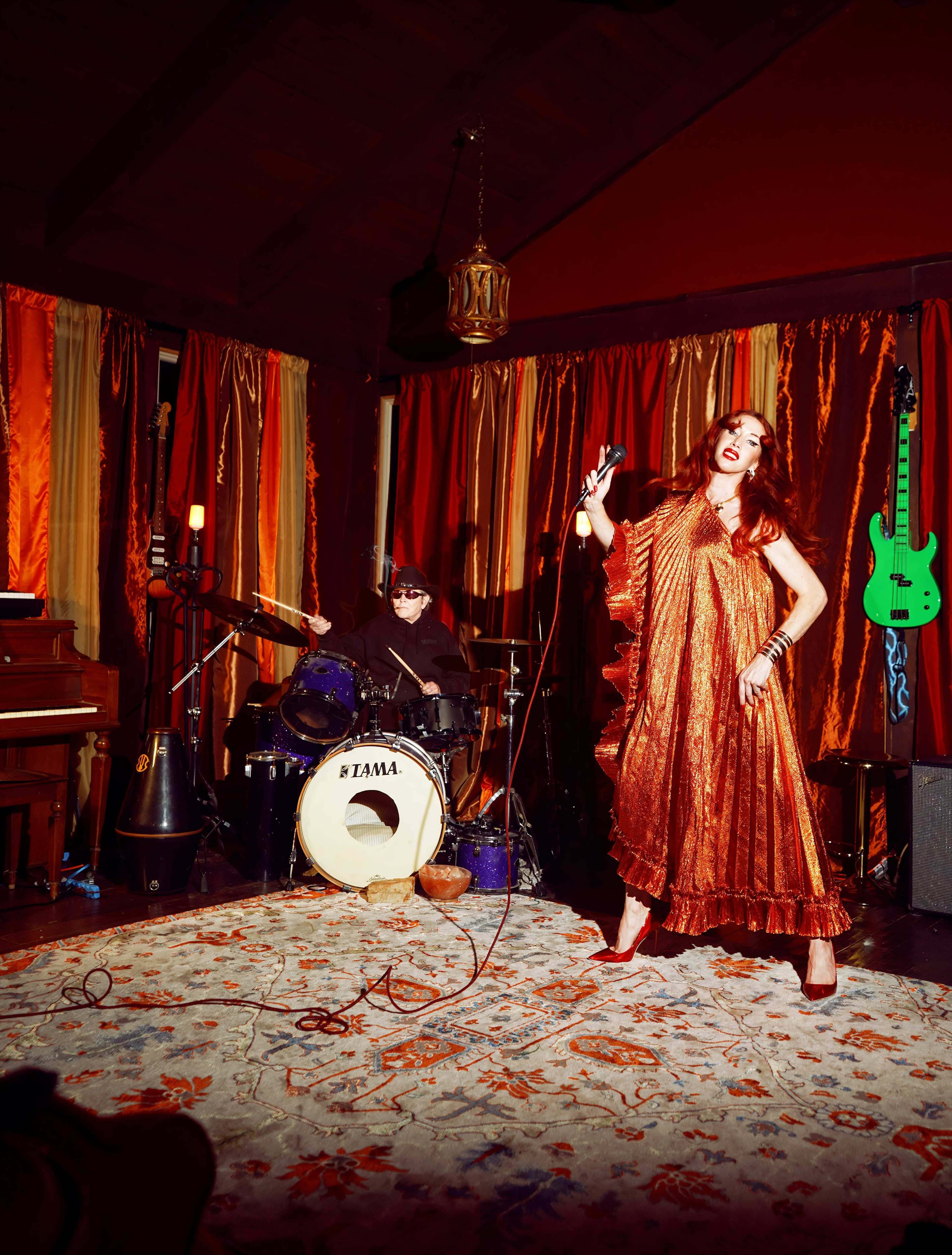 A singer in a flowing orange dress performs on stage while a drummer plays behind her in a venue decorated with vibrant curtains and a colorful rug.