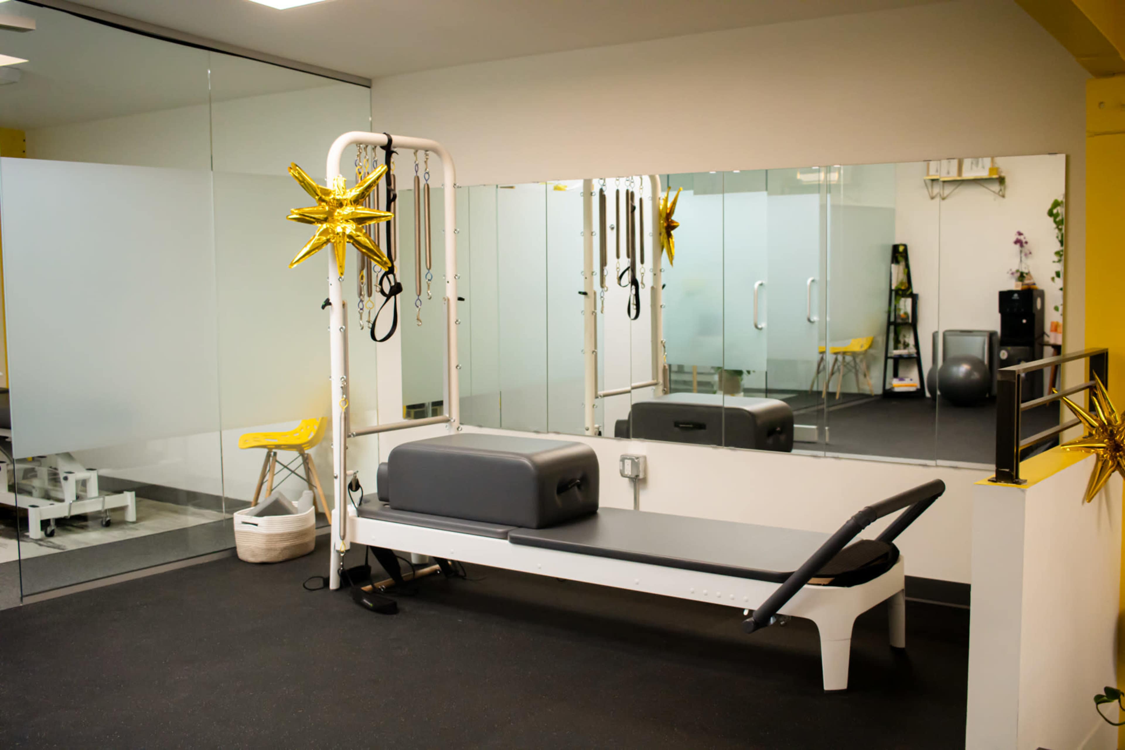 The image shows a Pilates studio featuring a reformer machine, a large mirror, and minimalistic decor with yellow accents.