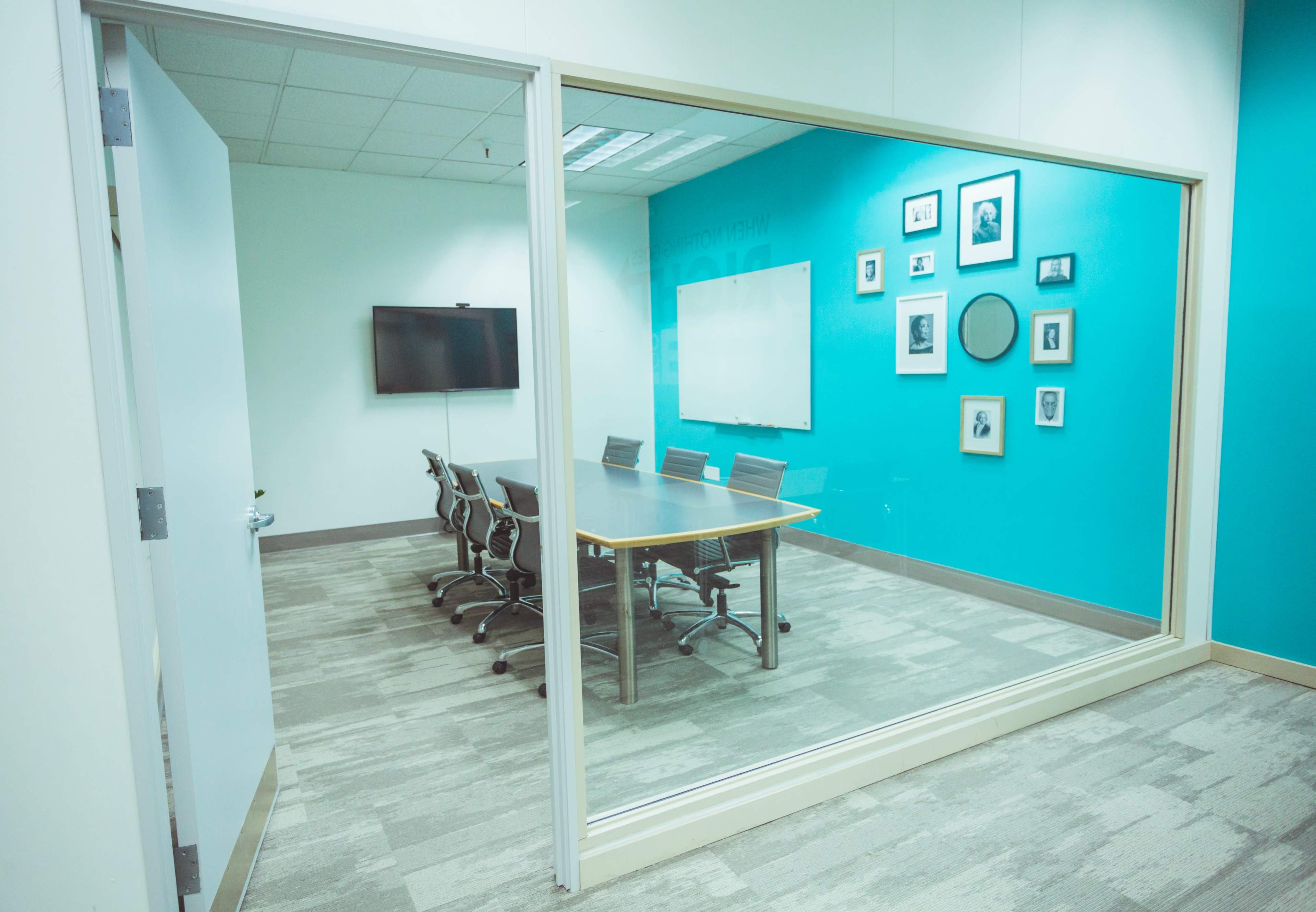 A glass-walled conference room features a long table surrounded by chairs and displays a series of framed pictures on a teal wall.
