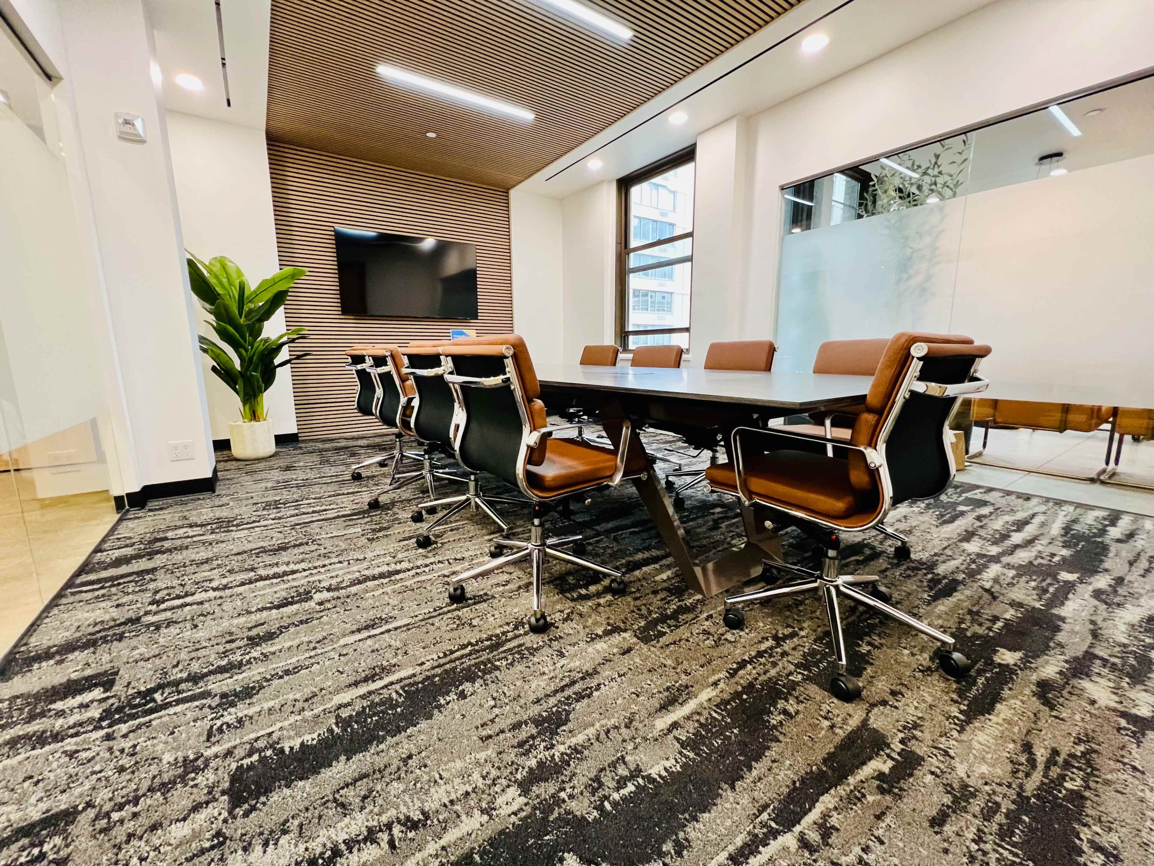 A modern conference room features a long table with rolling chairs, a large screen on the wall, and a plant in the corner.