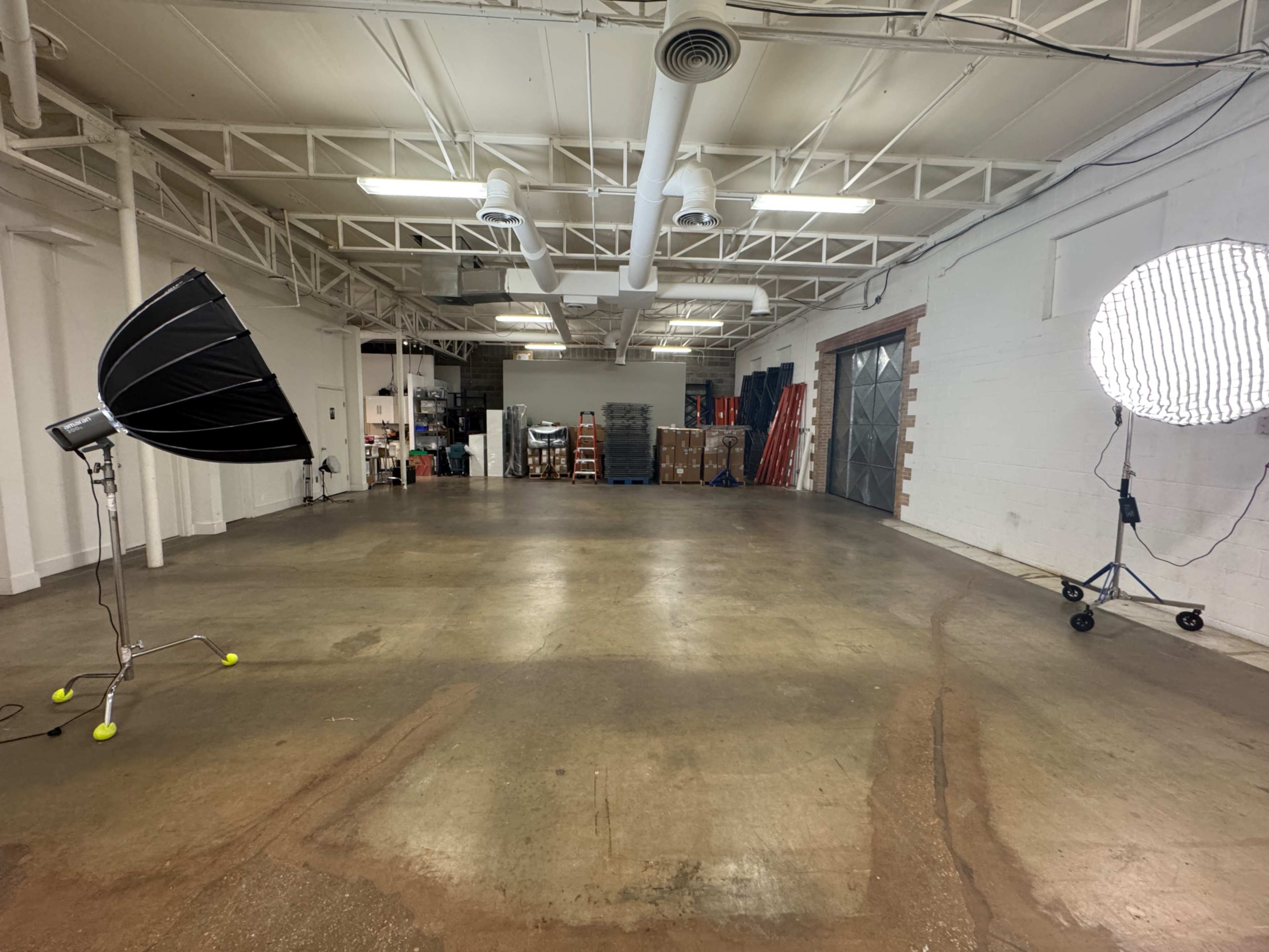 The image shows an empty industrial space with concrete floors, two large light sources on tripods, and stacks of equipment against the back wall.