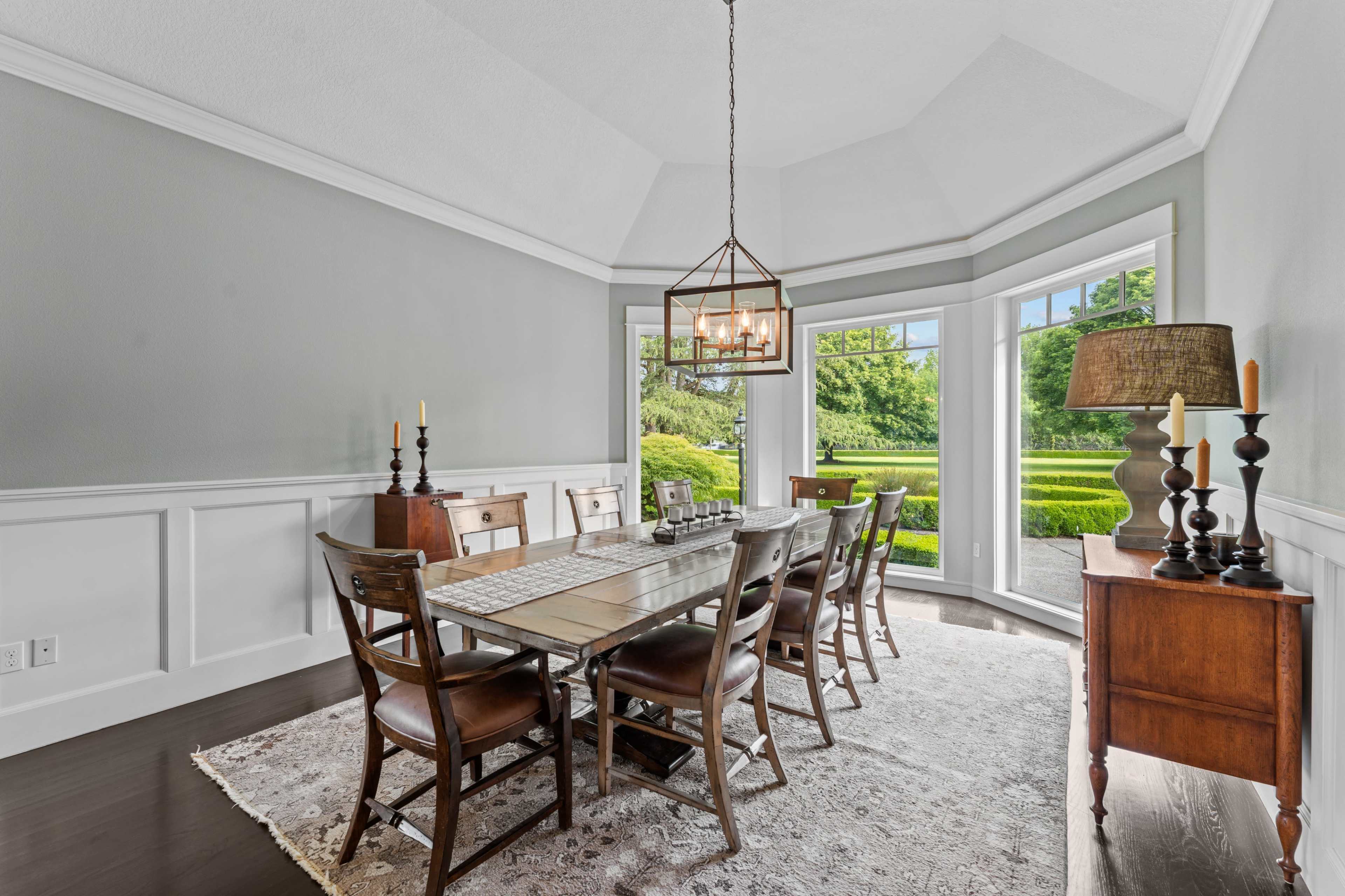 The image shows a well-lit dining room featuring a large wooden table surrounded by eight chairs, with windows offering a view of a landscaped garden.
