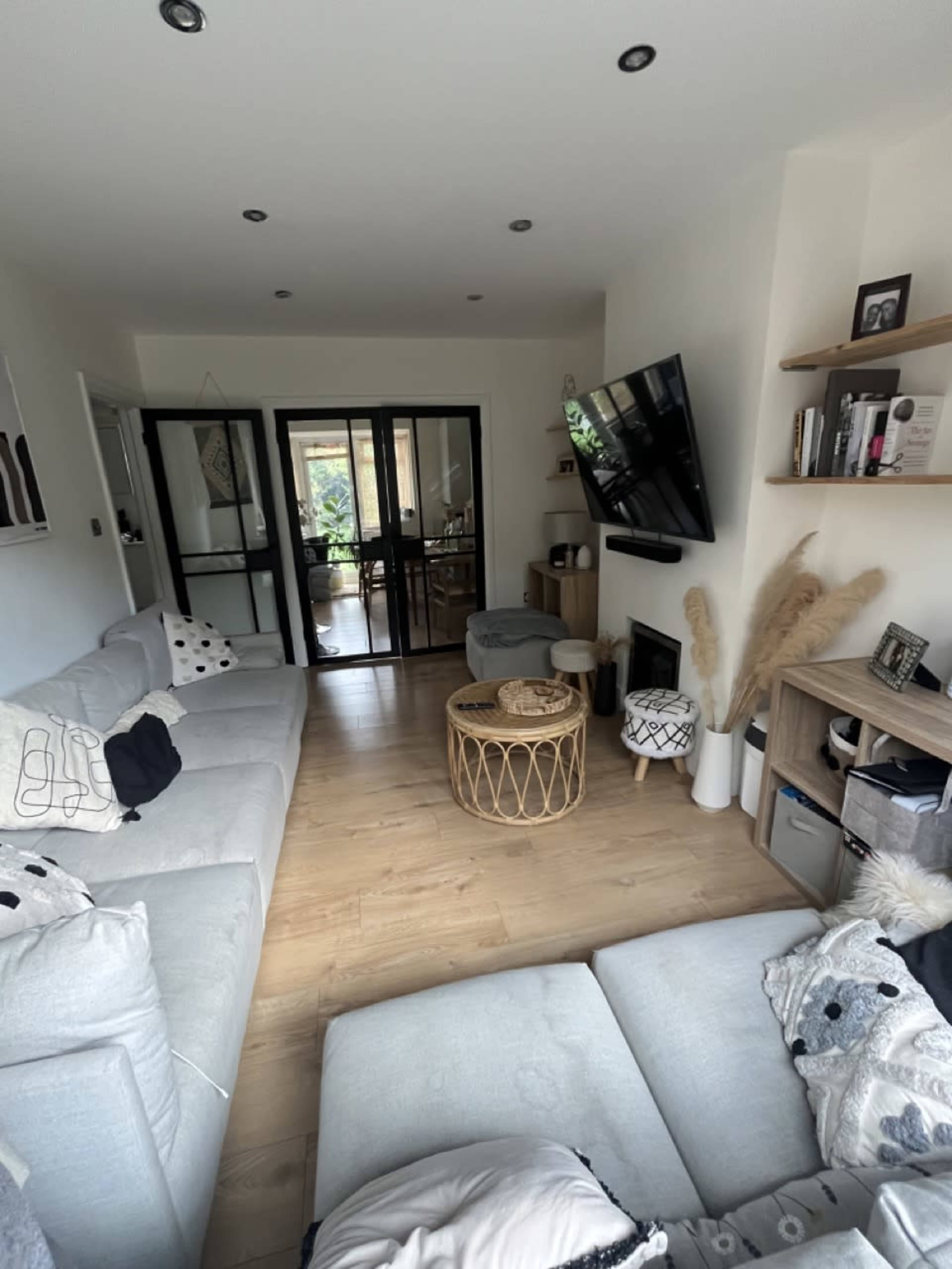 A modern living room features two light gray sofas arranged around a round wicker coffee table, with a television mounted on the wall and a small dining area visible through glass doors.