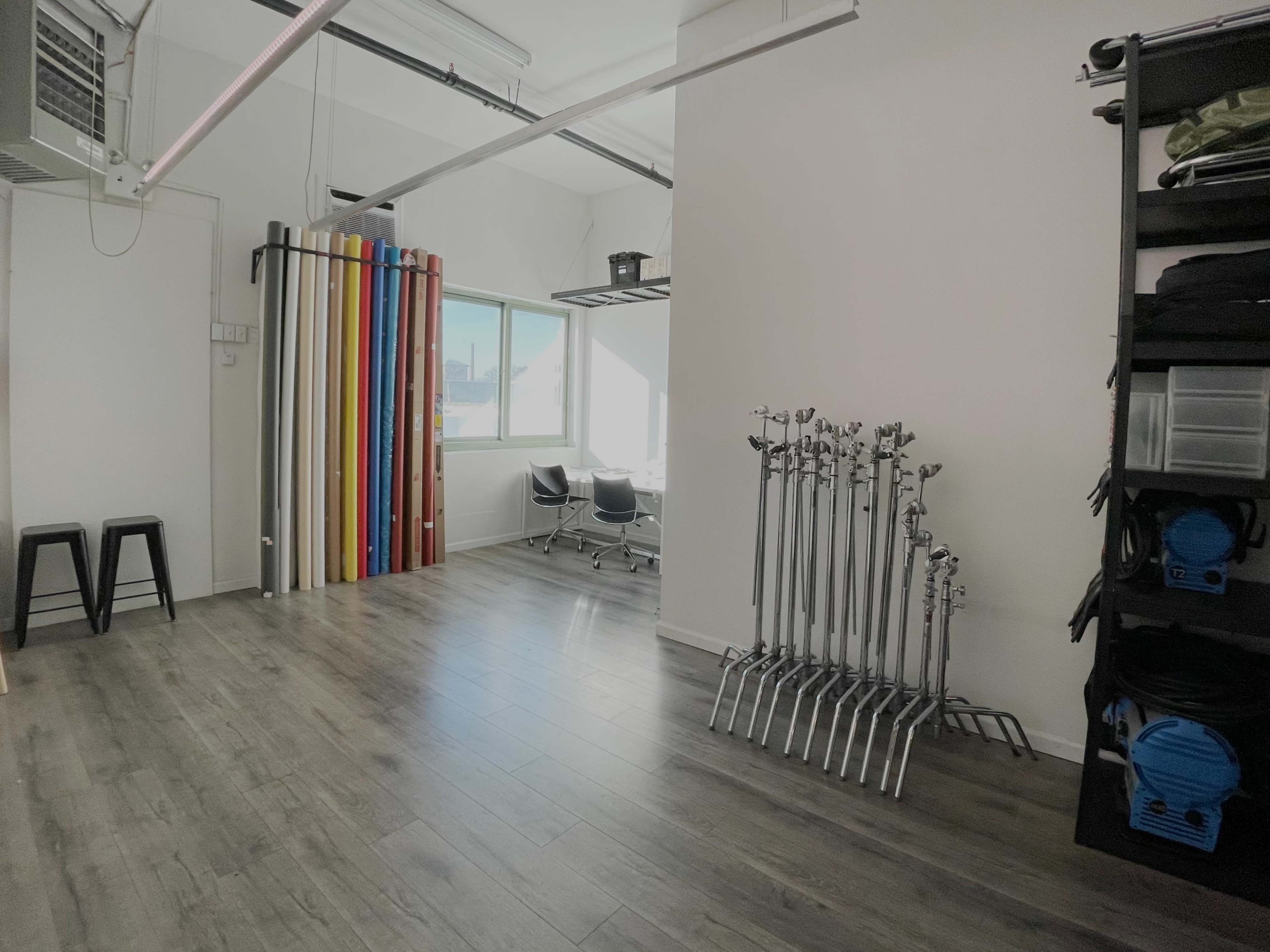 The image shows a spacious studio with colorful backdrop rolls and several light stands arranged on one side, adjacent to a workspace with two black chairs and a window.