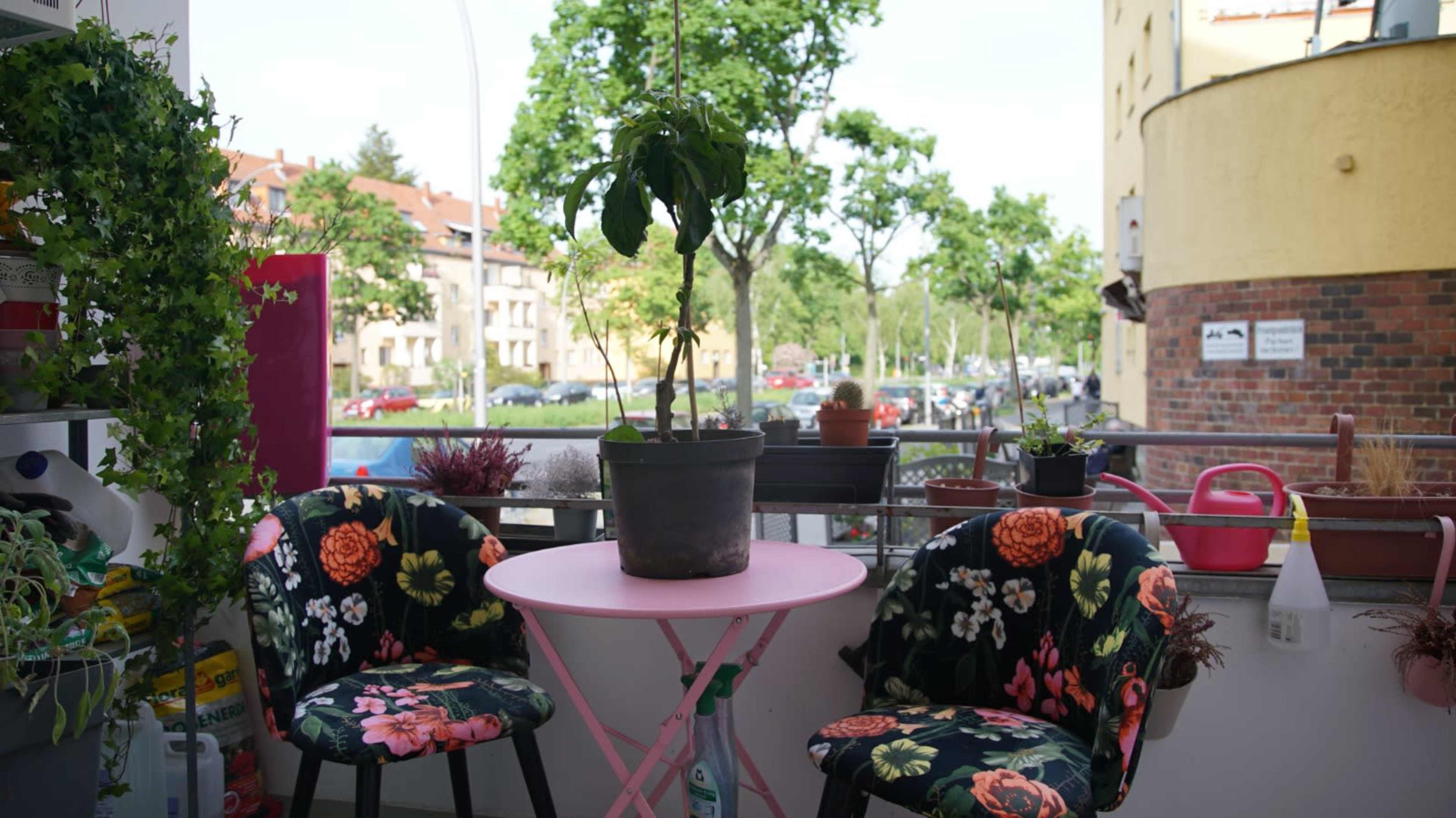 A small balcony features a round pink table, two floral-patterned chairs, and a potted plant, with a view of trees and buildings in the background.