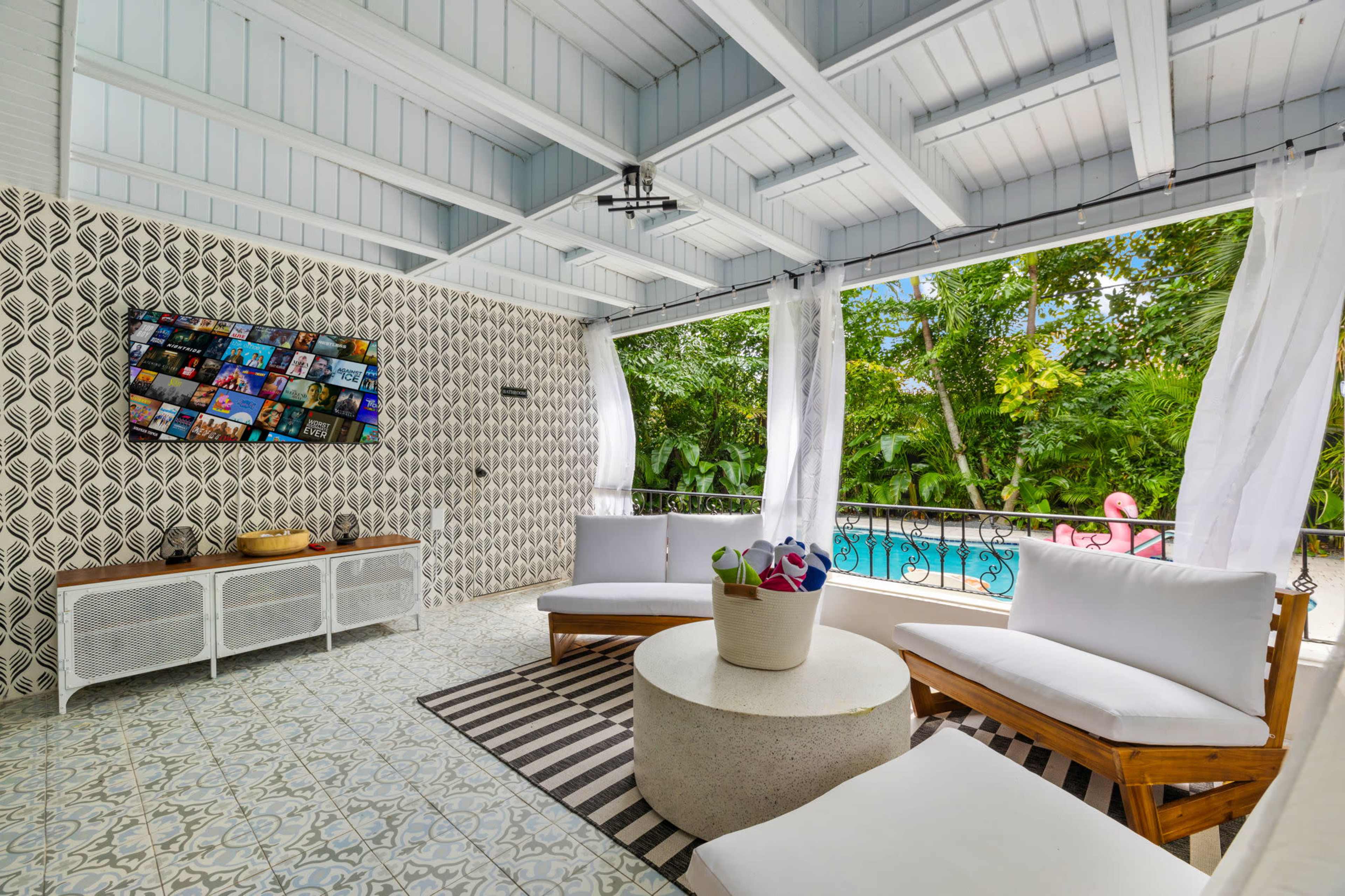 A modern outdoor lounge area features white sofas and a round table, with a television mounted on a patterned wall and tropical greenery visible through open curtains.