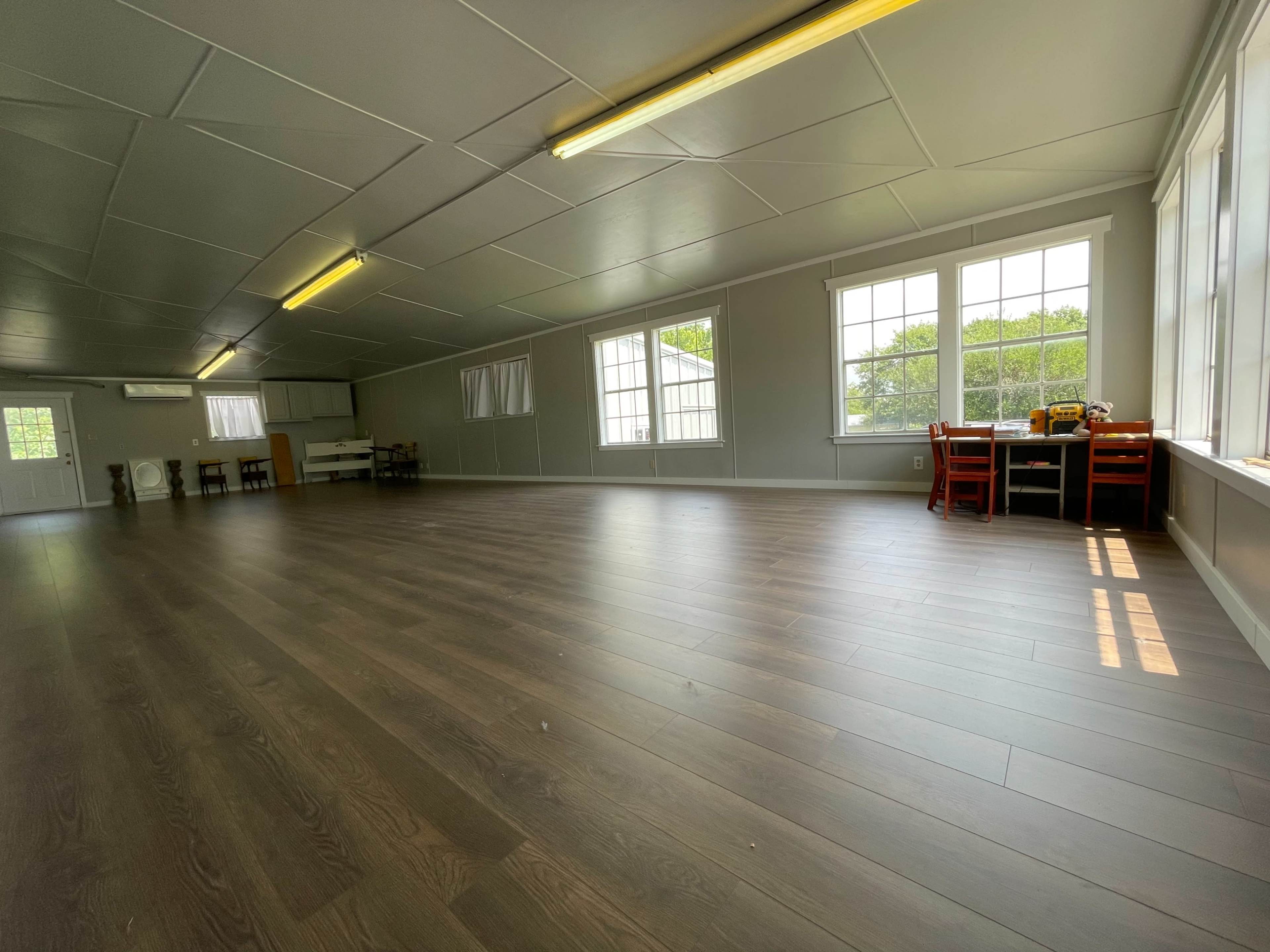 A spacious, empty room with wooden flooring and large windows along one side, furnished with a few tables and chairs.