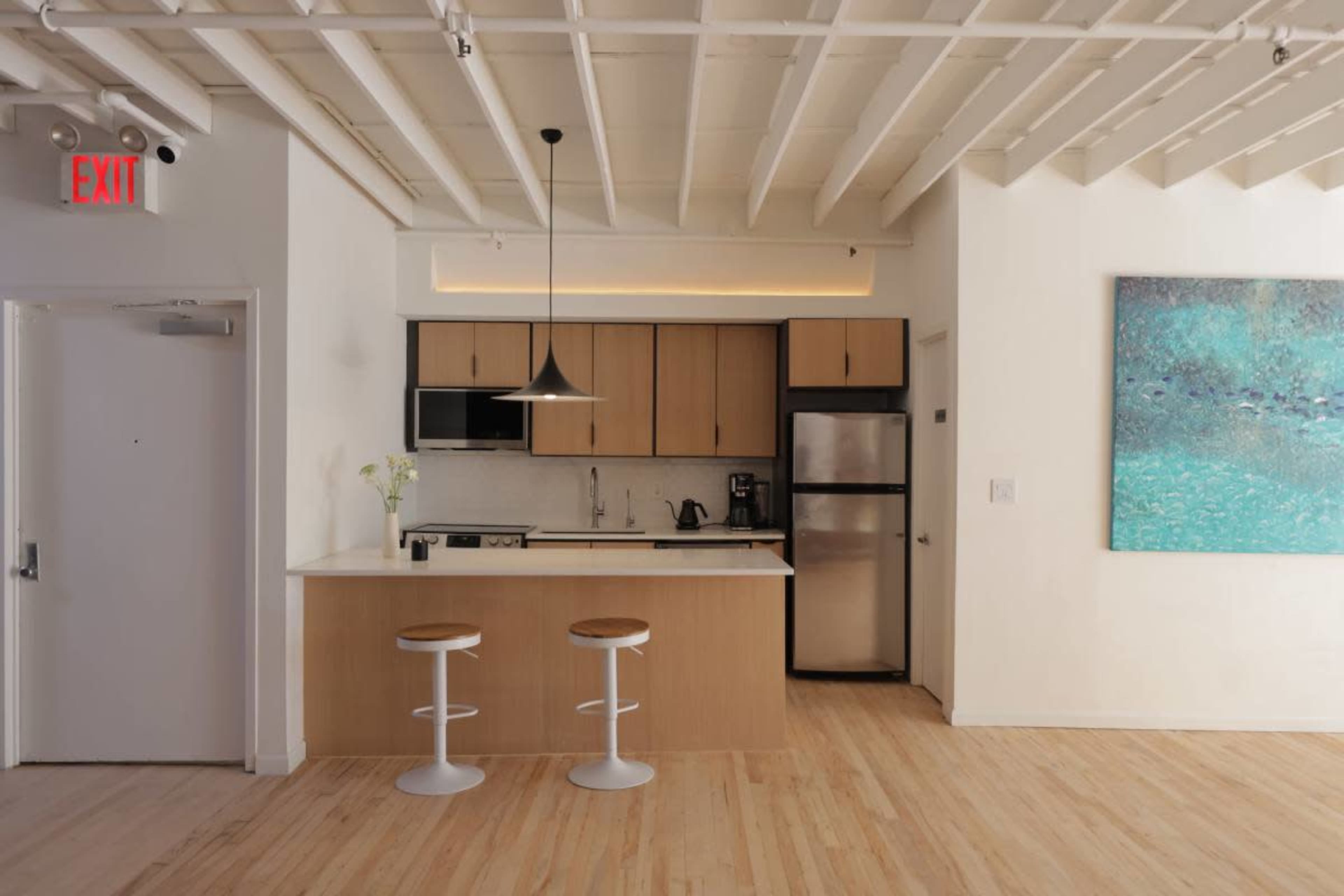 The image shows a modern kitchen with a wooden island, two white stools, and a refrigerator, accompanied by a large piece of artwork on the wall.