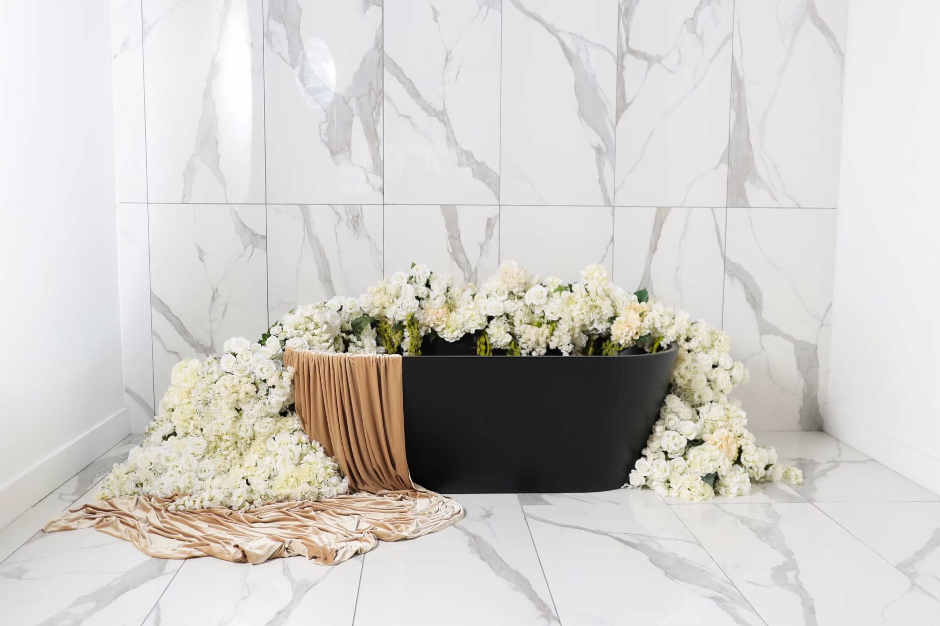 A black bathtub is surrounded by an array of white flowers against a marble wall.