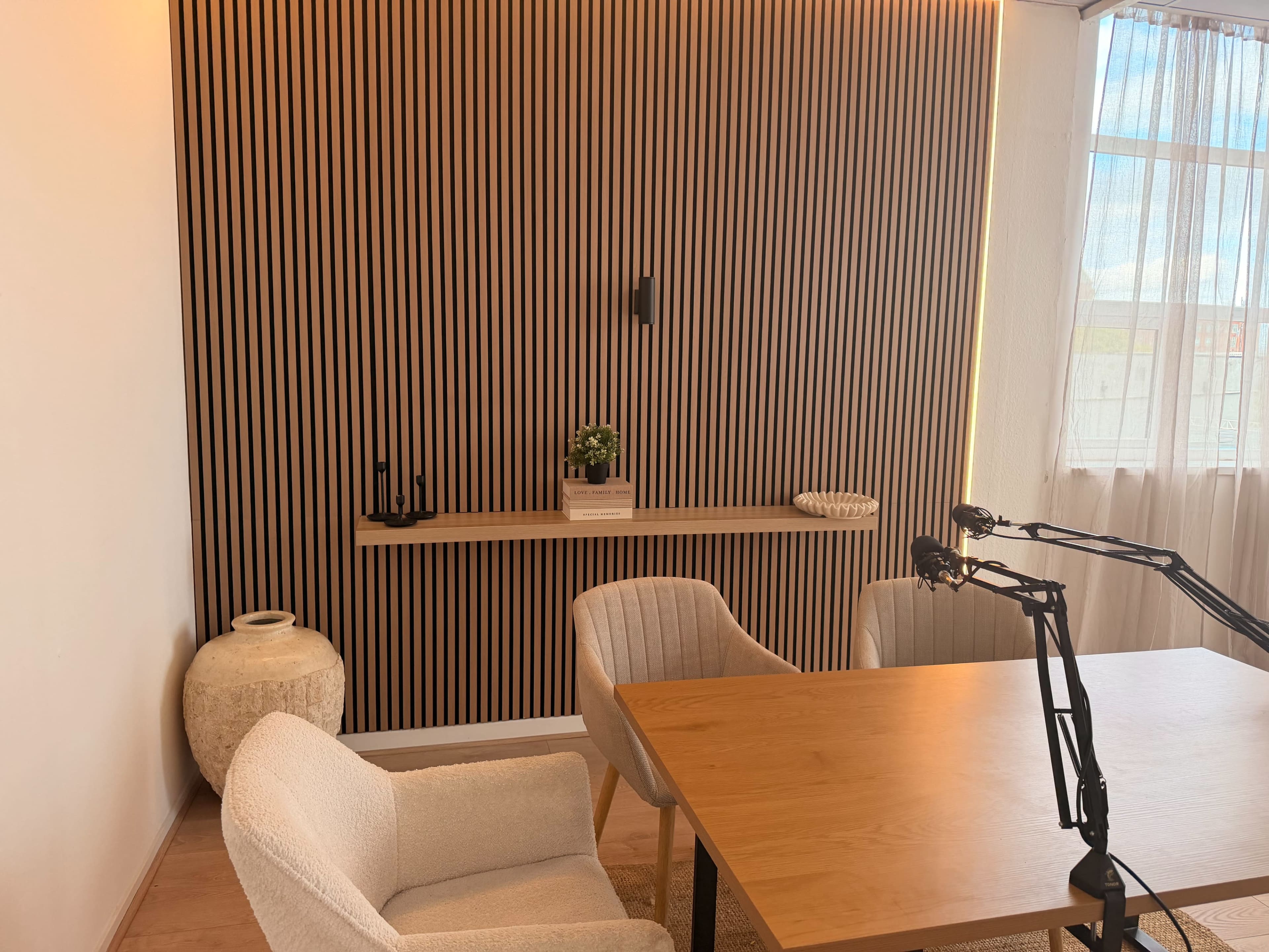 The image shows a modern, minimalistic room featuring a wooden table, two upholstered chairs, and a striped wooden accent wall, with natural light coming through a large window.