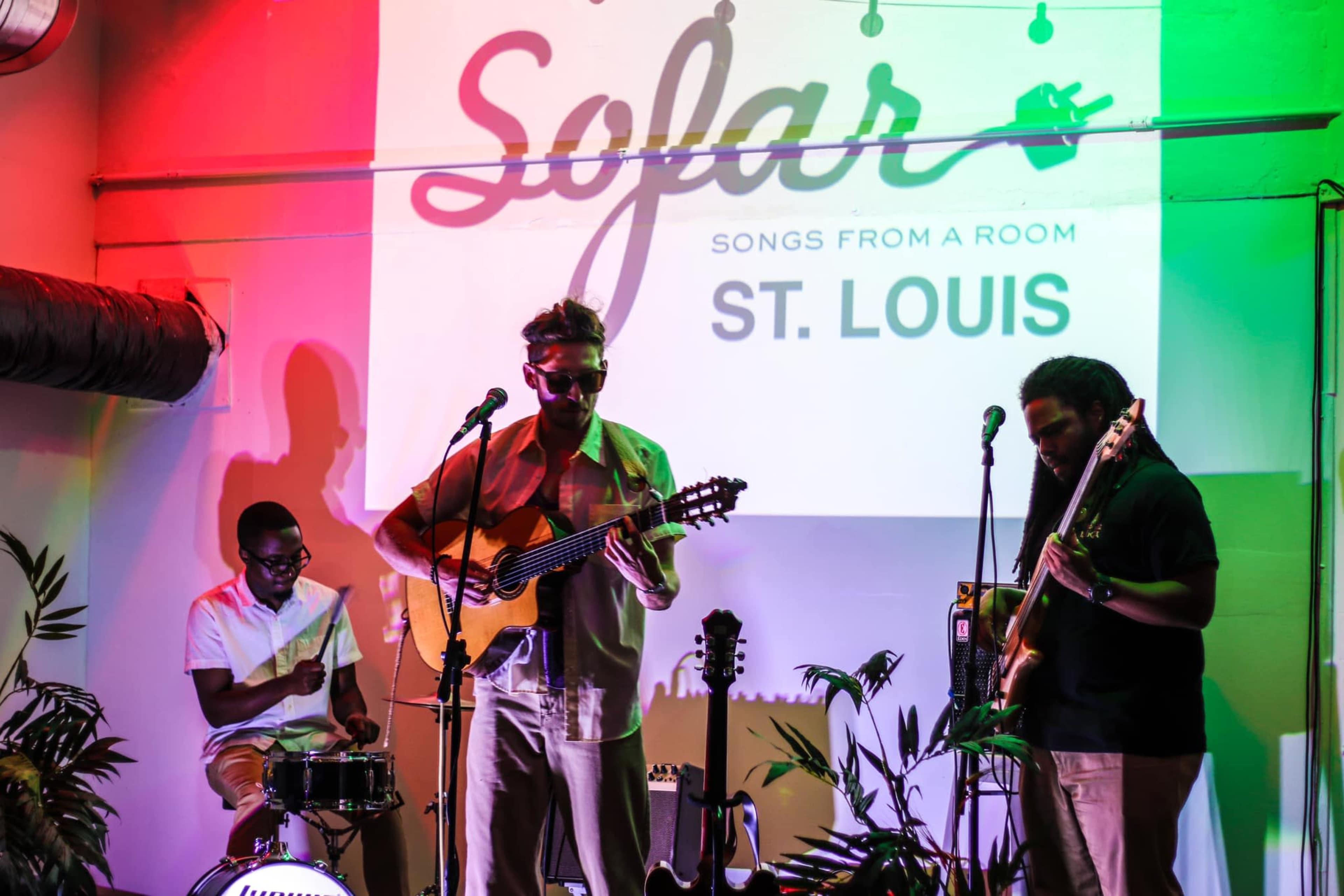 A musician plays an acoustic guitar while another musician stands with a bass guitar and a drummer is seated behind a drum set during a Sofar concert in St. Louis.
