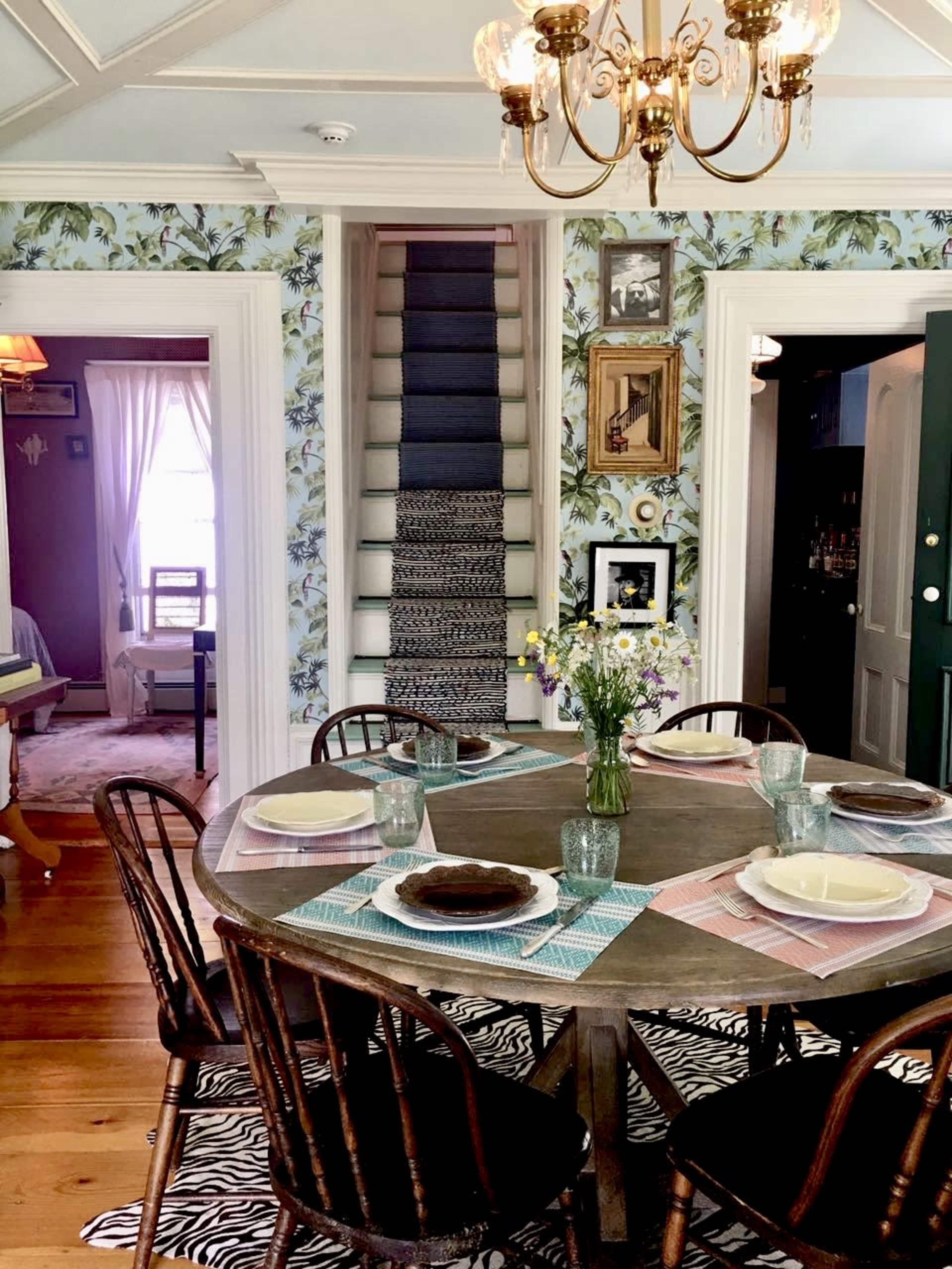 A round wooden dining table is set with plates and glasses in a patterned dining room, flanked by a staircase and floral wallpaper.