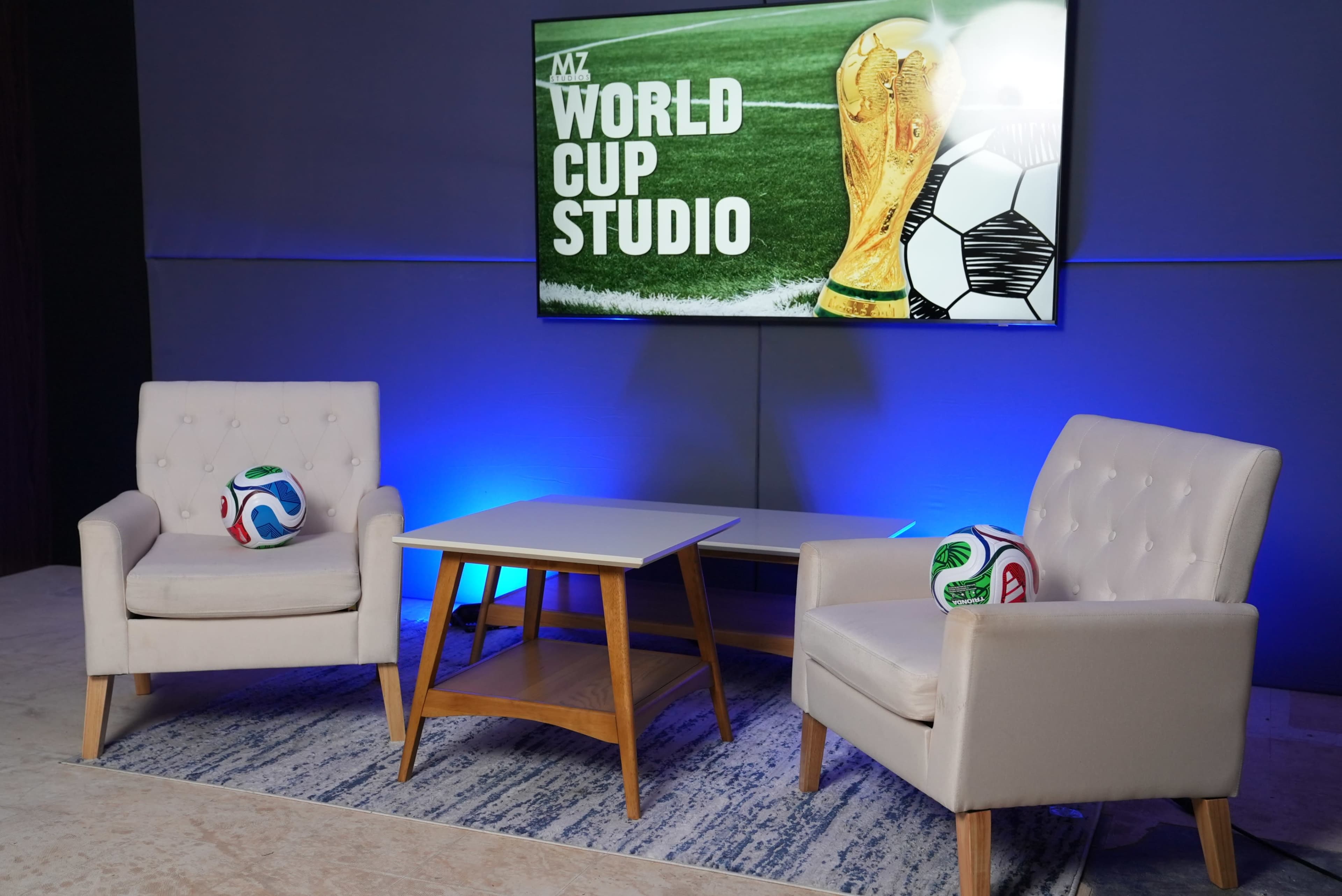 The image shows a modern studio setup with two white chairs and a wooden table, featuring a television screen displaying a World Cup graphic along with soccer balls on the chairs.