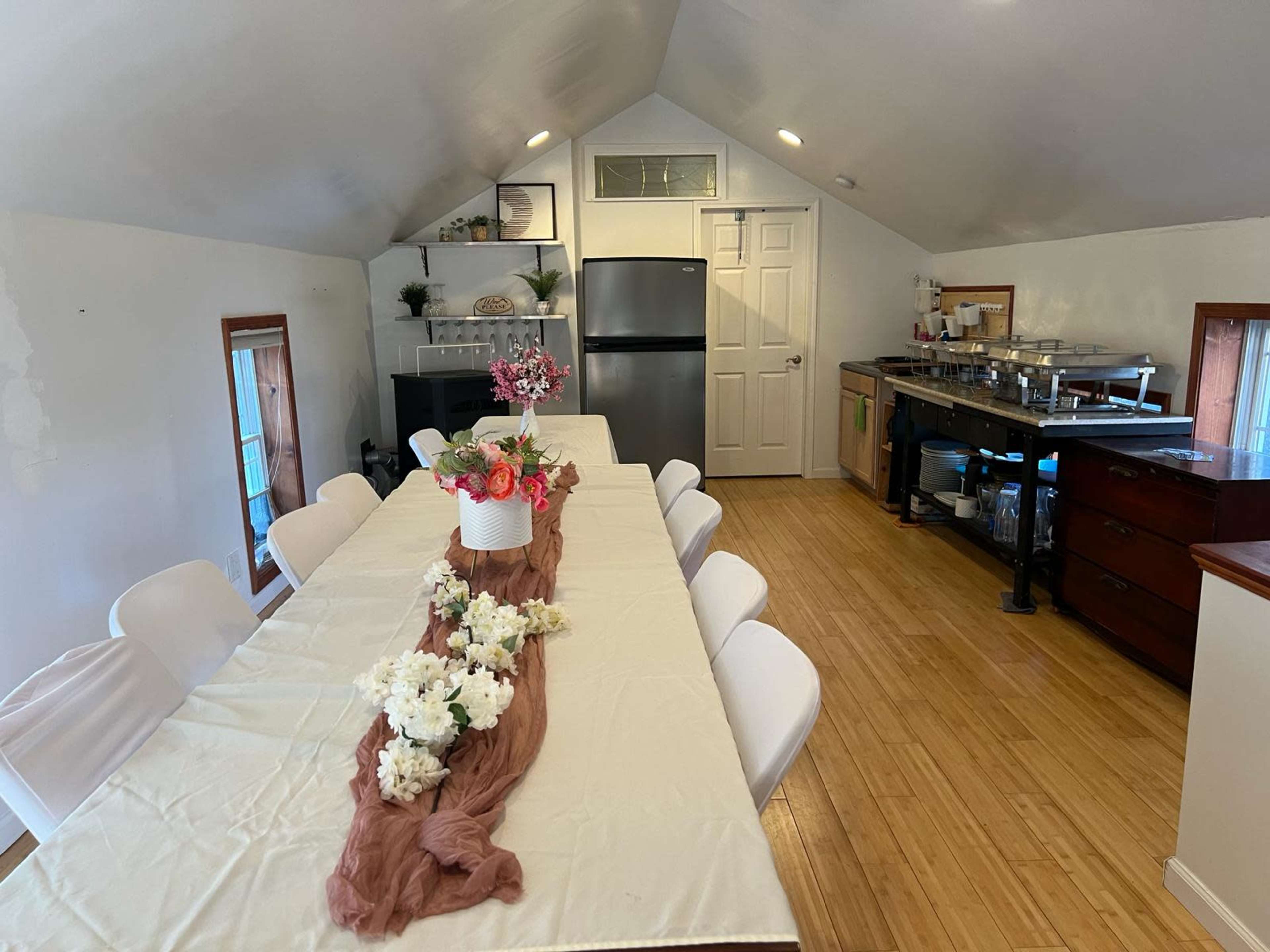 A spacious, well-lit kitchen and dining area featuring a long table with a tablecloth, surrounded by white chairs and decorated with flowers, and a kitchenette with modern appliances.