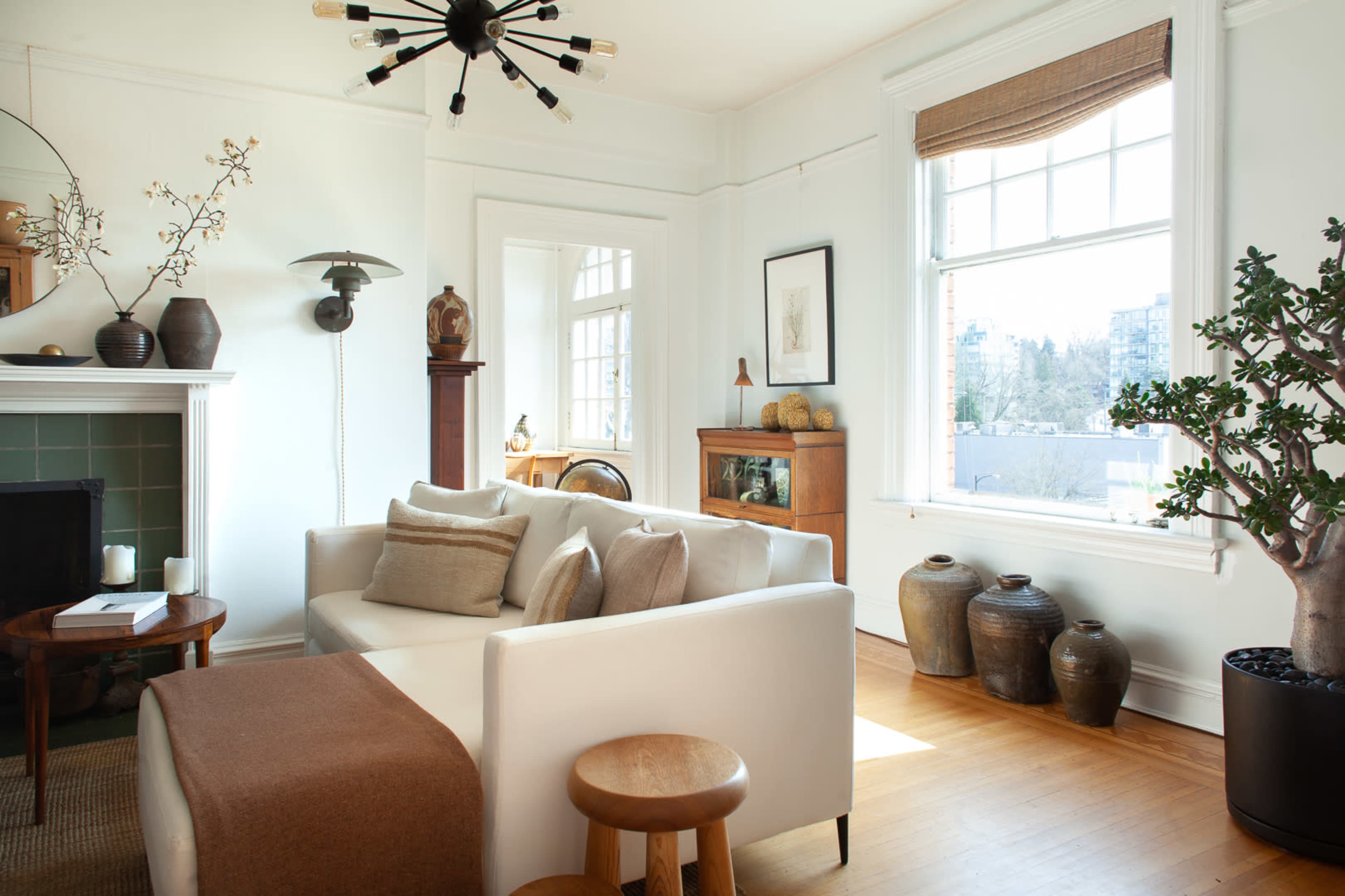 The image shows a bright living room featuring a white sofa, wooden stools, decorative plants, and a large window with a view.