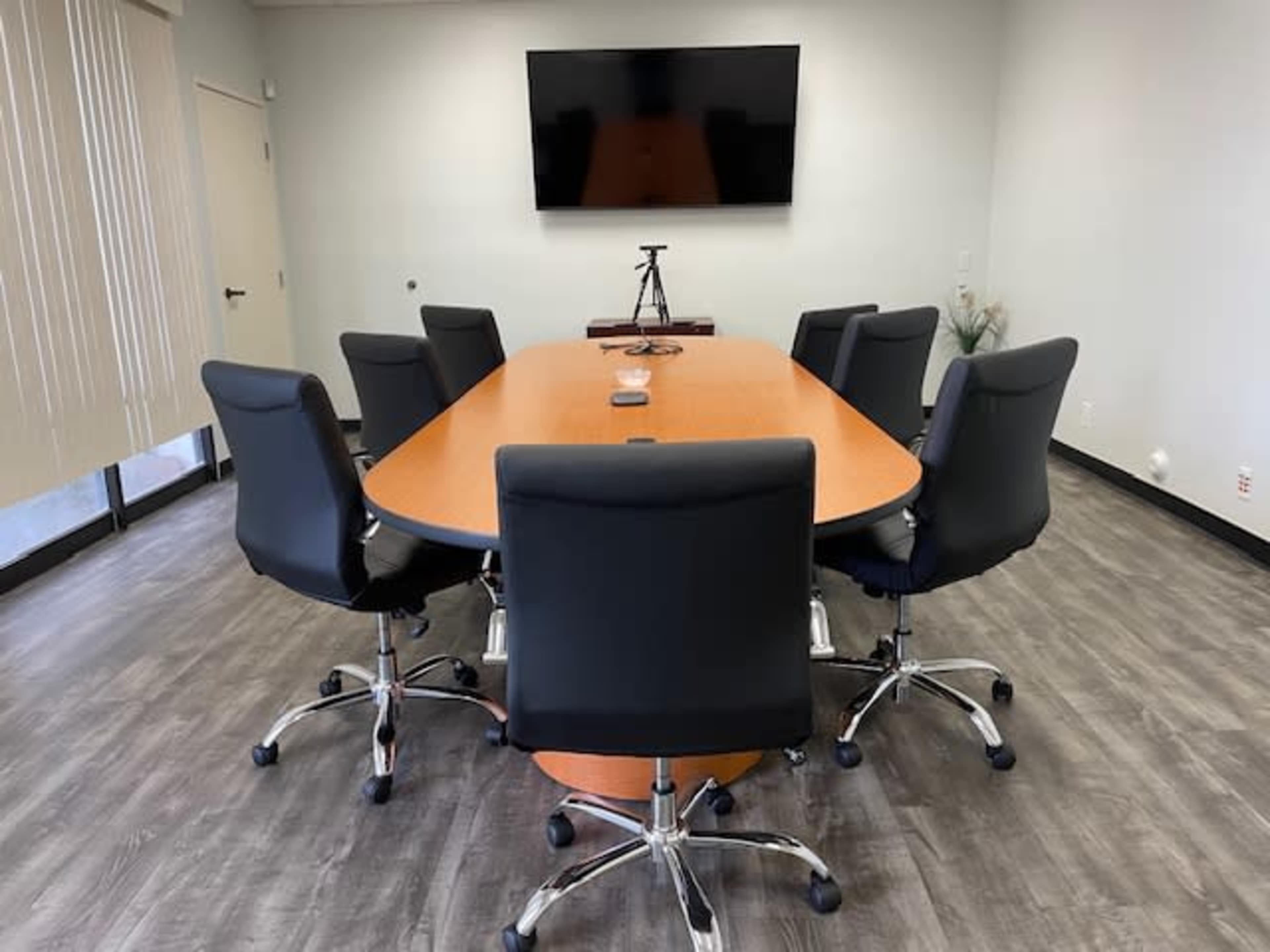 A conference room features a large oval table surrounded by six black office chairs, with a wall-mounted screen at one end.