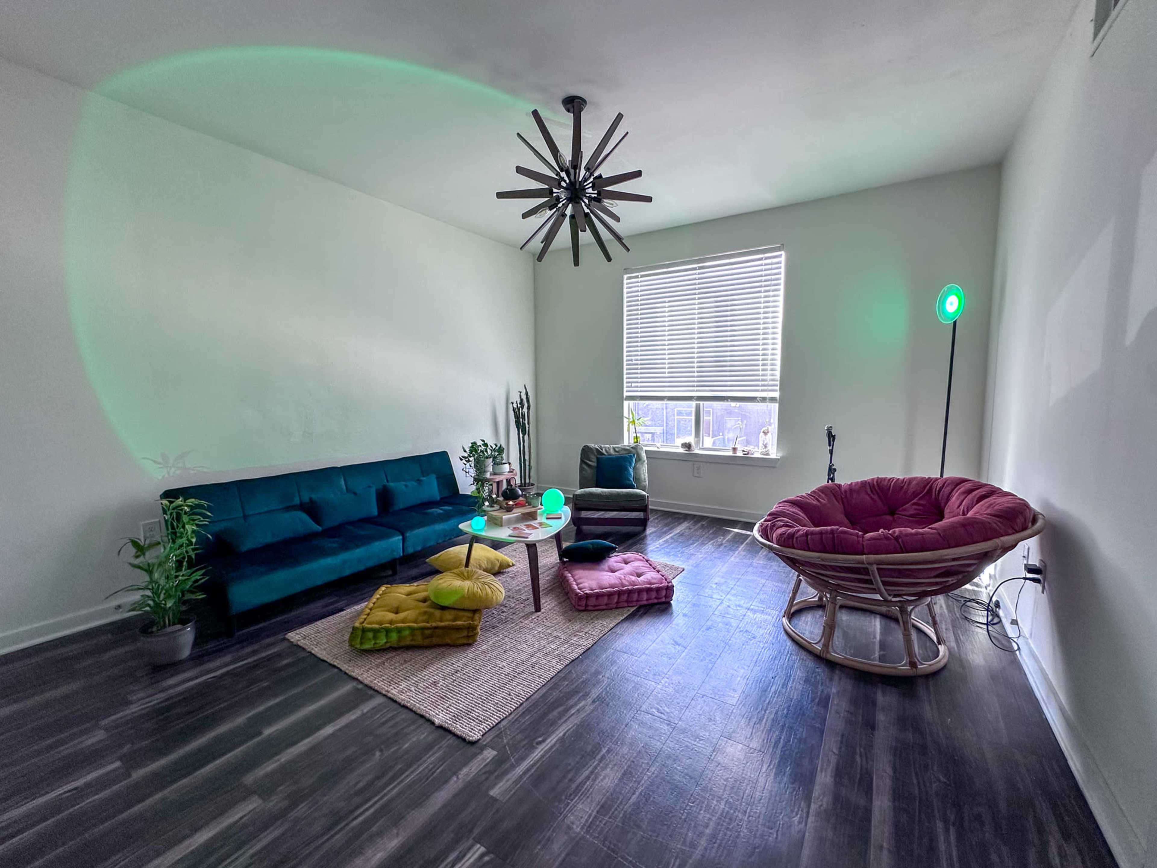 The image shows a modern living room featuring a dark green sofa, a round pink chair, various cushions, a small table, and a green light source near the window.