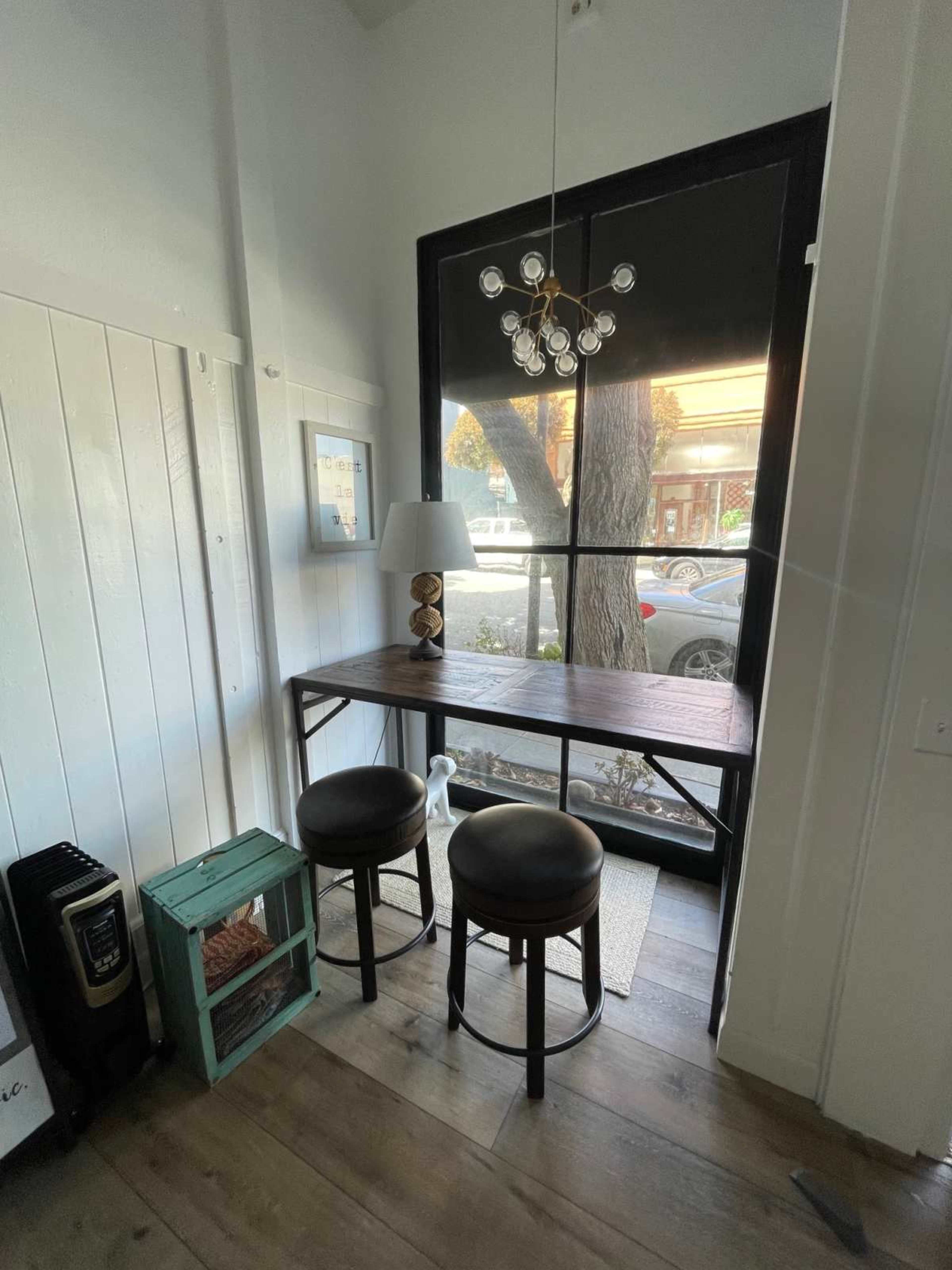 A small seating area with two black stools and a wooden table next to a large window, featuring a lamp and a decorative cabinet.