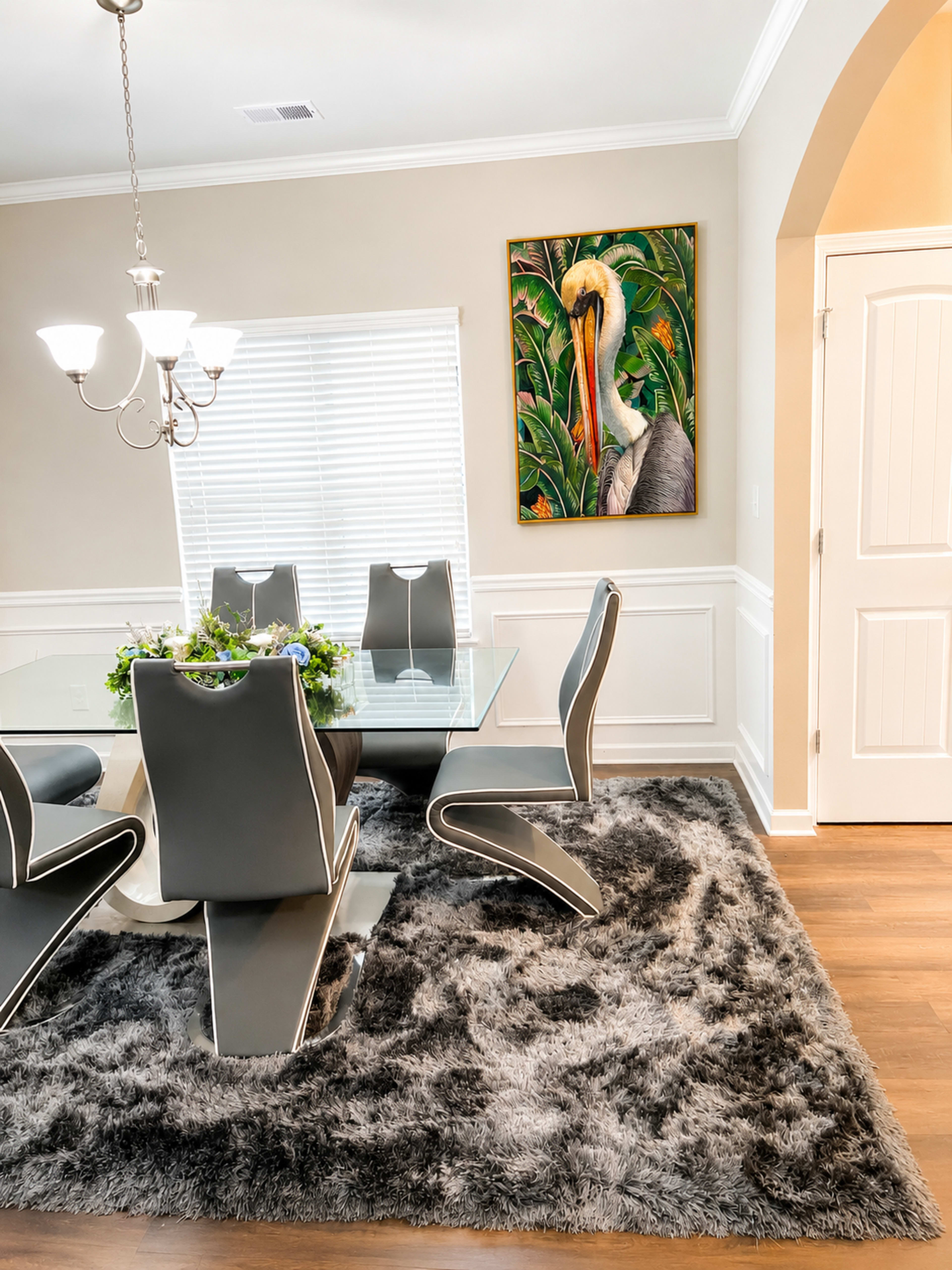 The image shows a modern dining room featuring a glass table surrounded by stylish chairs, a large textured rug, and a vibrant painting of a bird on the wall.