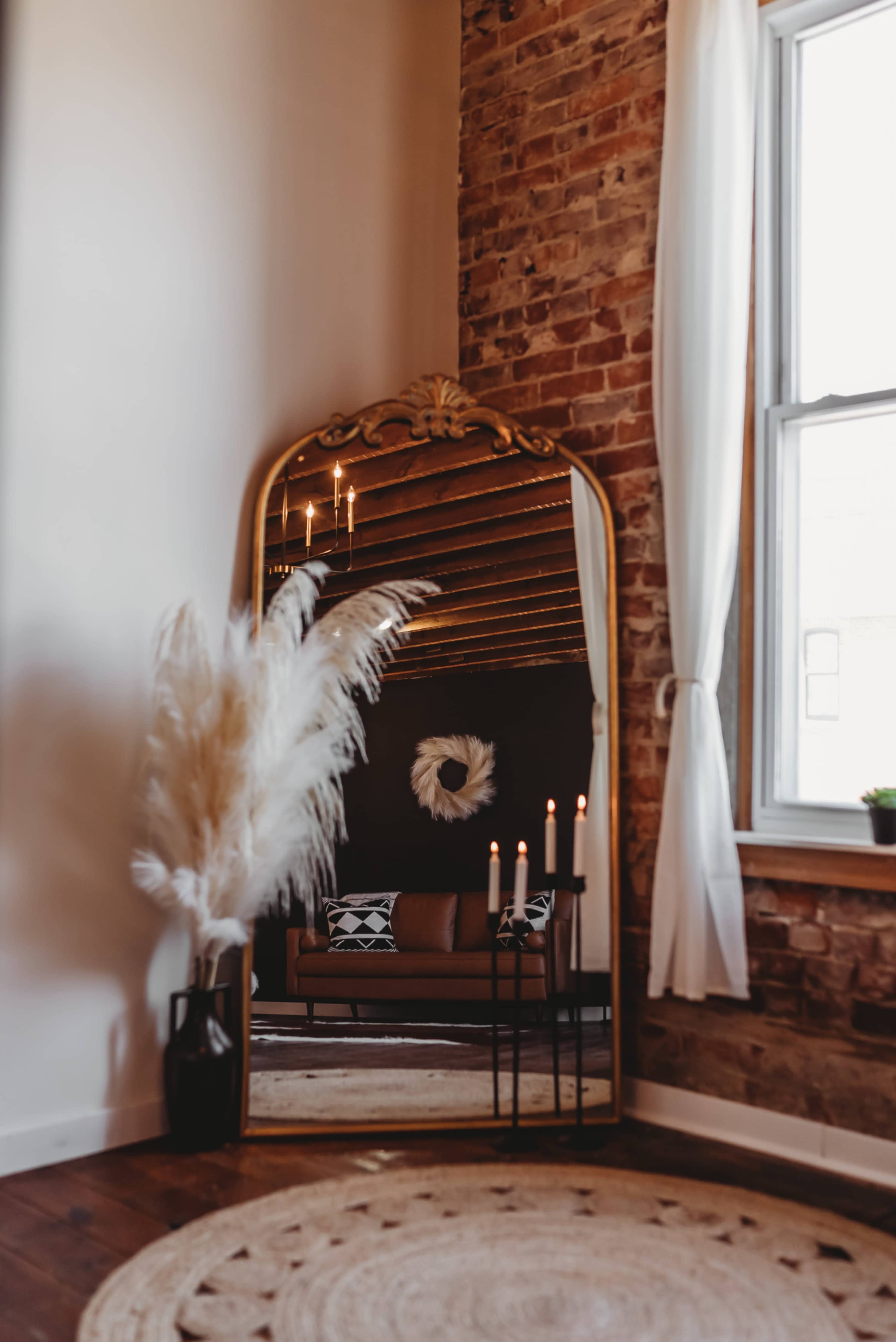 A tall, ornate mirror reflects a cozy seating area with a plant and candles in a warmly lit room featuring exposed brick walls.